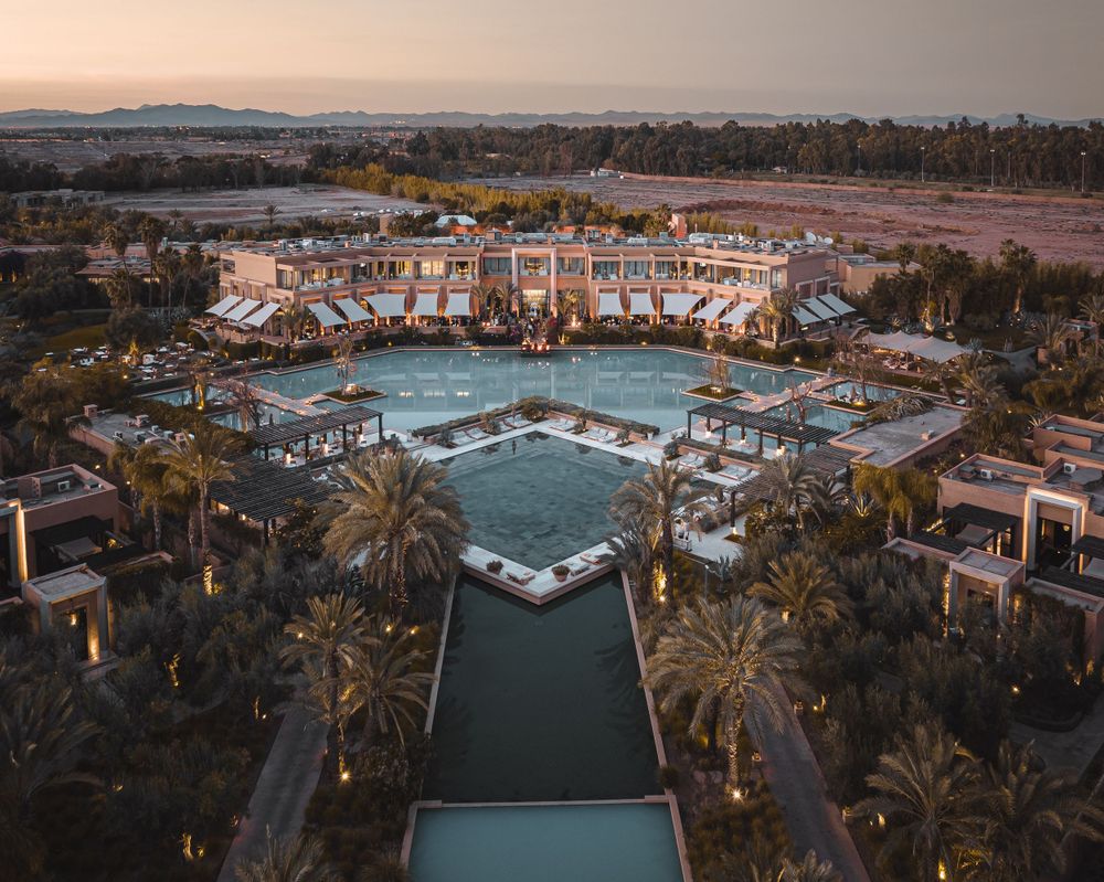 Aerial view of a Mediterranean-style resort complex with pools and palm trees during sunset.