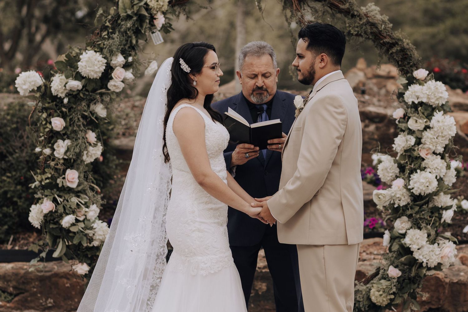 A wedding ceremony takes place under a floral arch with white roses in an outdoor garden setting.