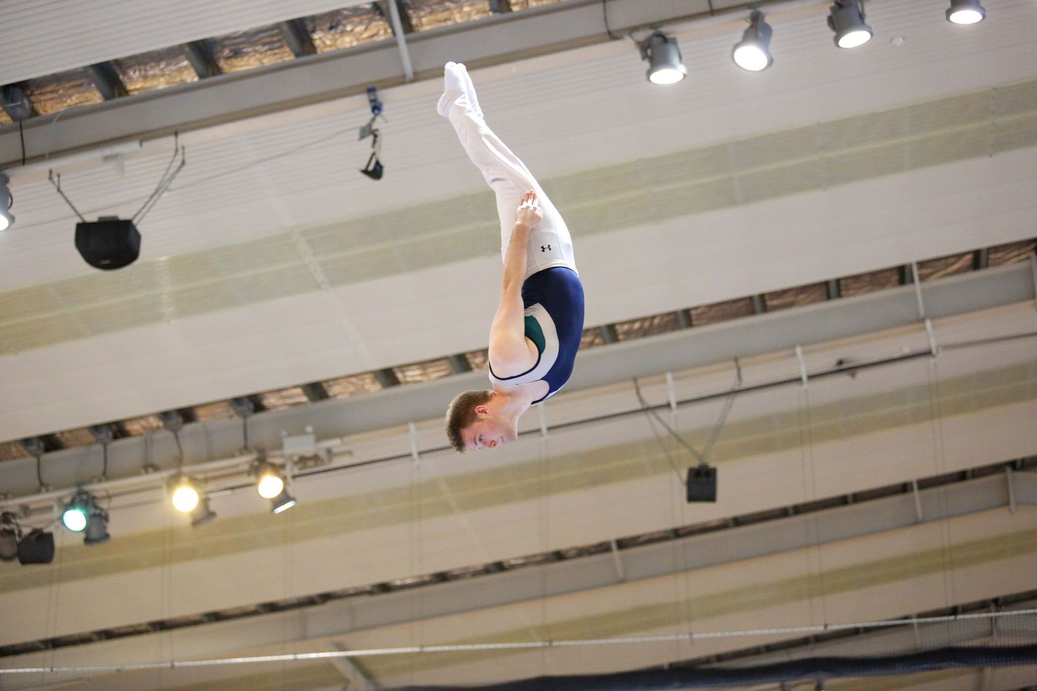 Gymnast executes aerial maneuver against ceiling spotlights in indoor arena.