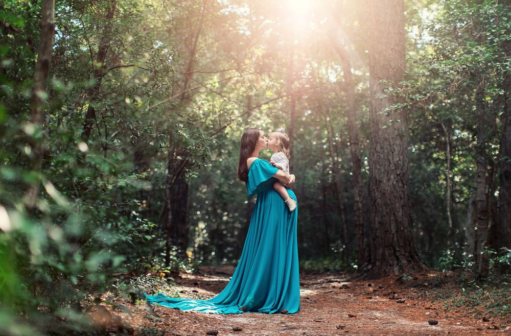 Woman in flowing teal dress stands in sunlit forest clearing.