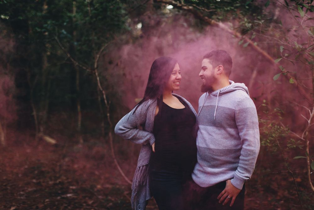 Couple shares intimate moment surrounded by pink smoke in forest setting.