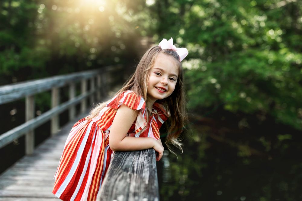 Young child in red and gray dress smiling on wooden bridge with sunlight and water in background.