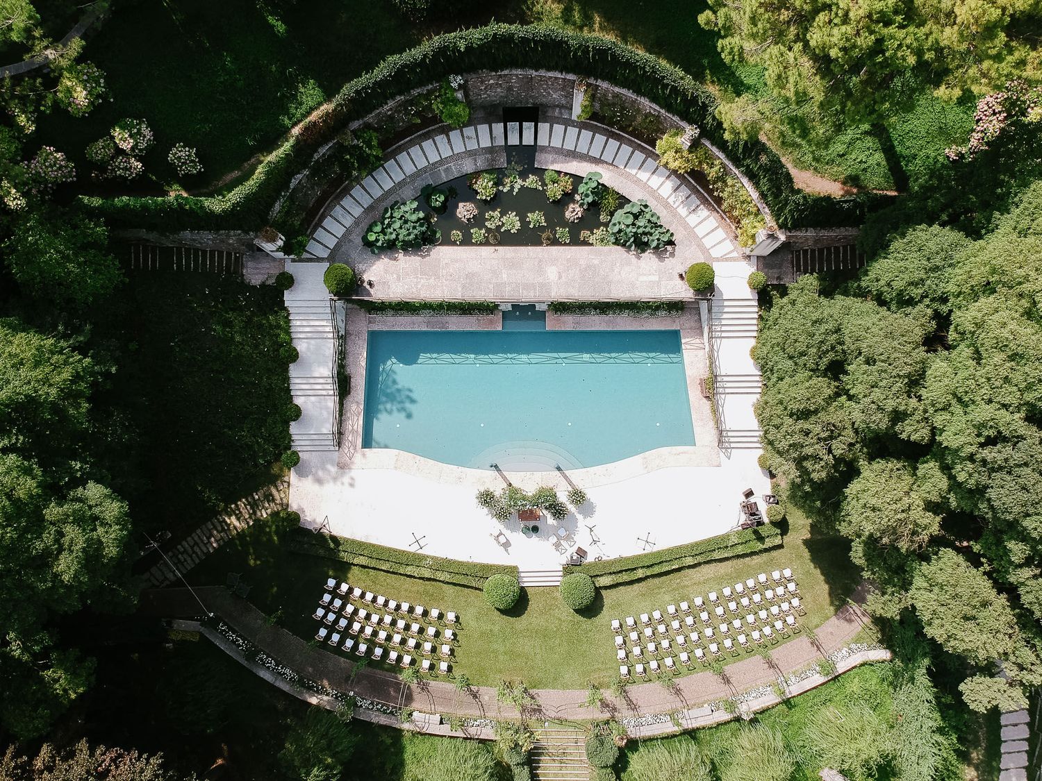 Aerial view of an elegant swimming pool surrounded by lush gardens with a classical arched colonnade and tiered seating.