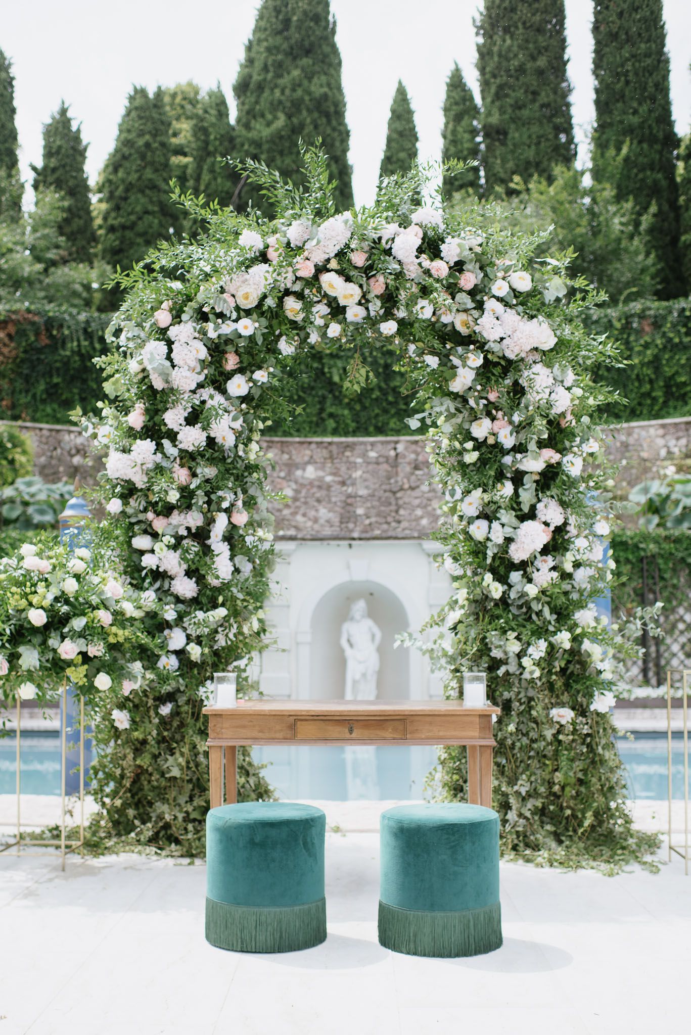 Floral wedding arch with white roses and greenery over turquoise poufs by a pool.