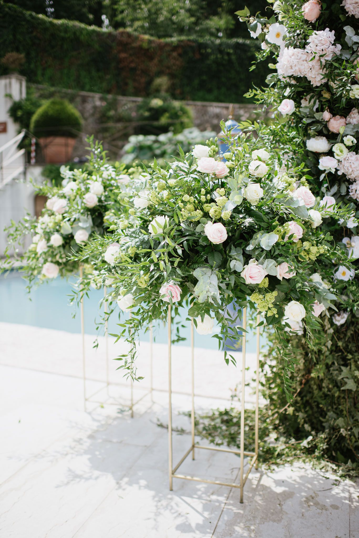 Elegant garden wedding decor with lush white and pink floral arrangements on gold stands against greenery.