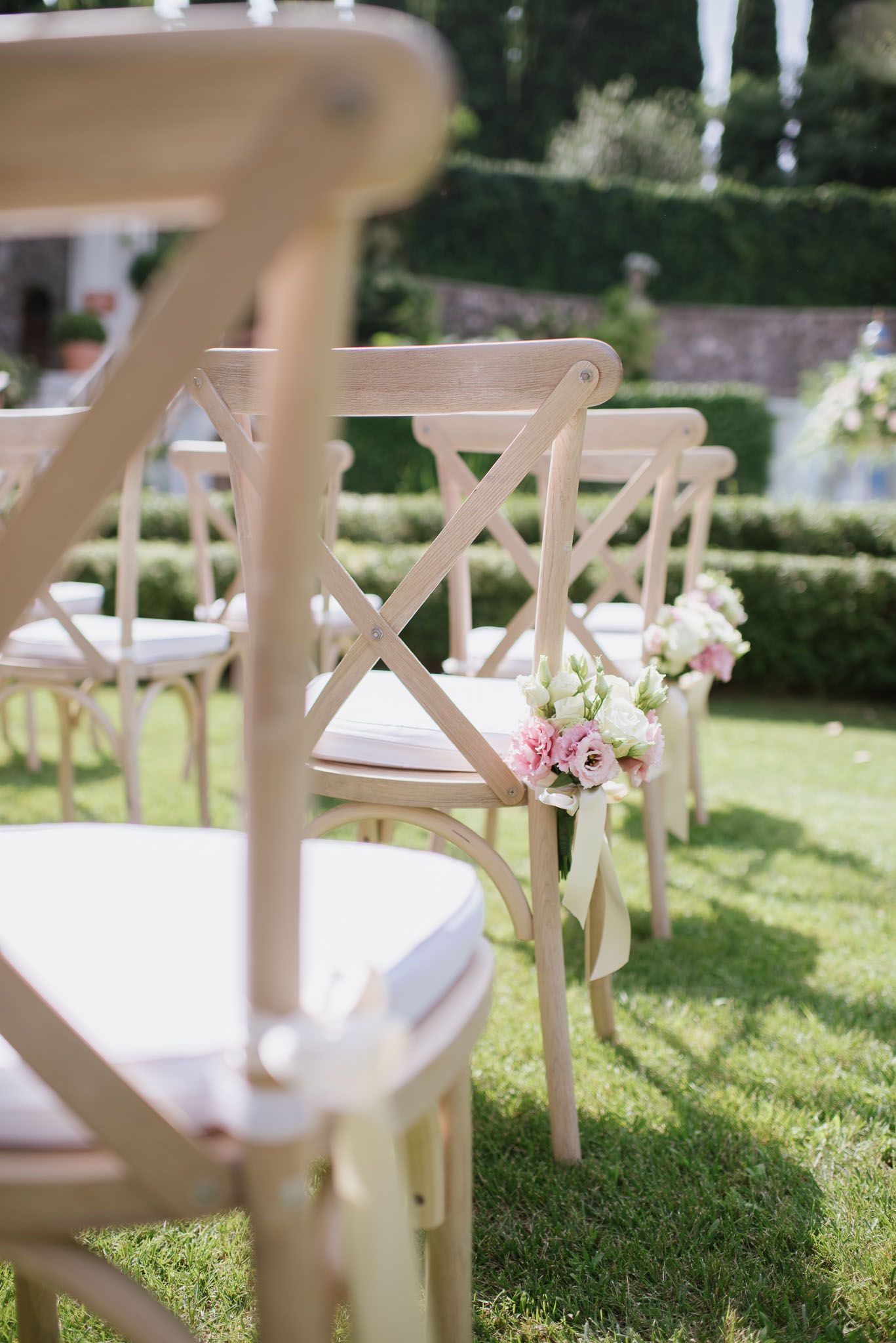 Cross-back wooden ceremony chairs decorated with pink floral arrangements in garden setting.