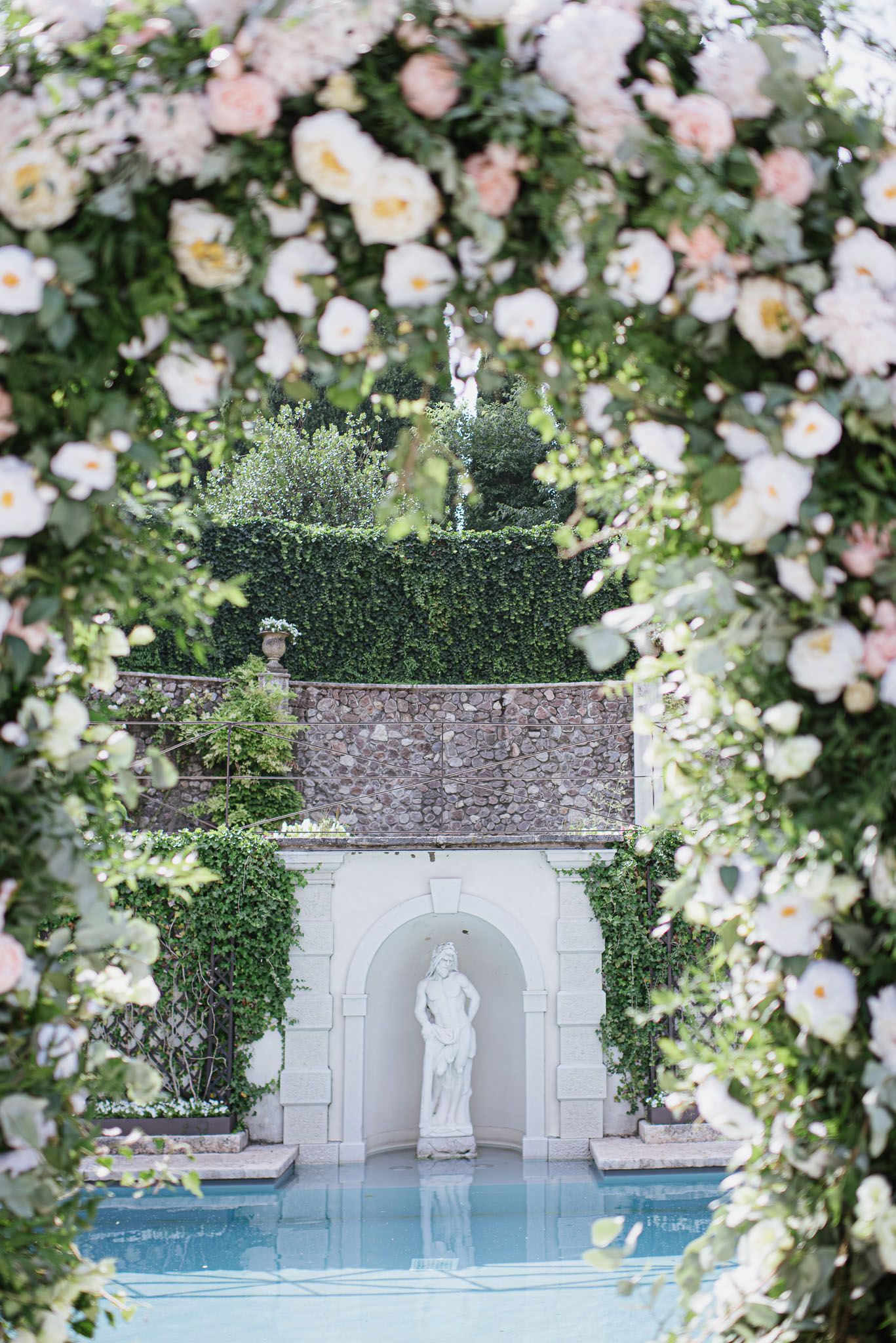 Romantic pool area framed by pink and white rose arch with classical statue in stone alcove.