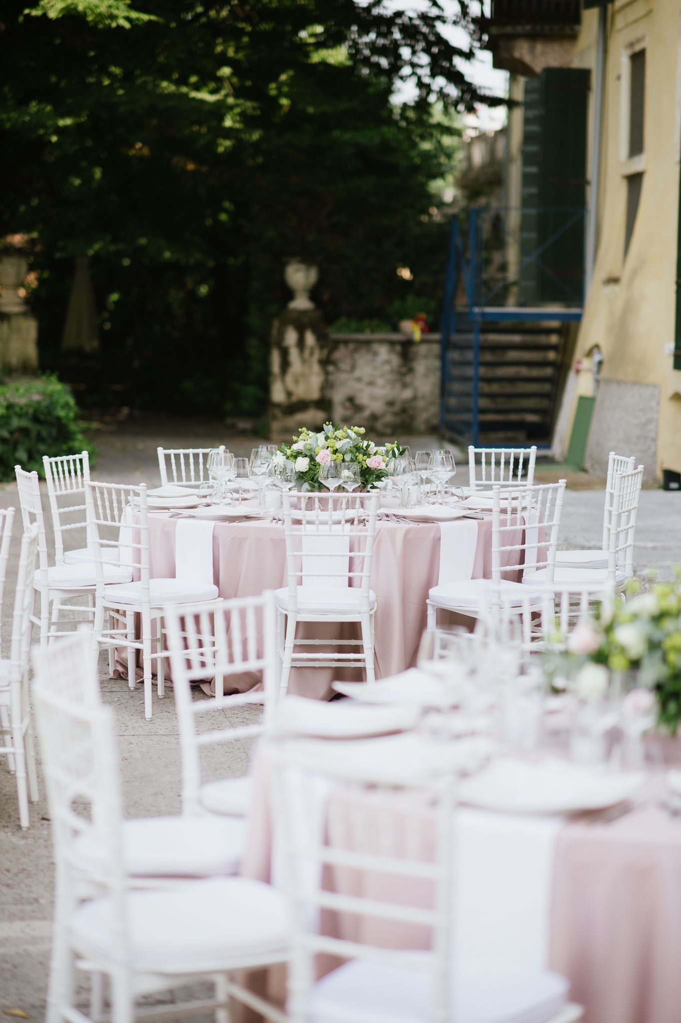 Elegant outdoor wedding reception setup with white chiavari chairs and soft pink tablecloths in a garden setting.