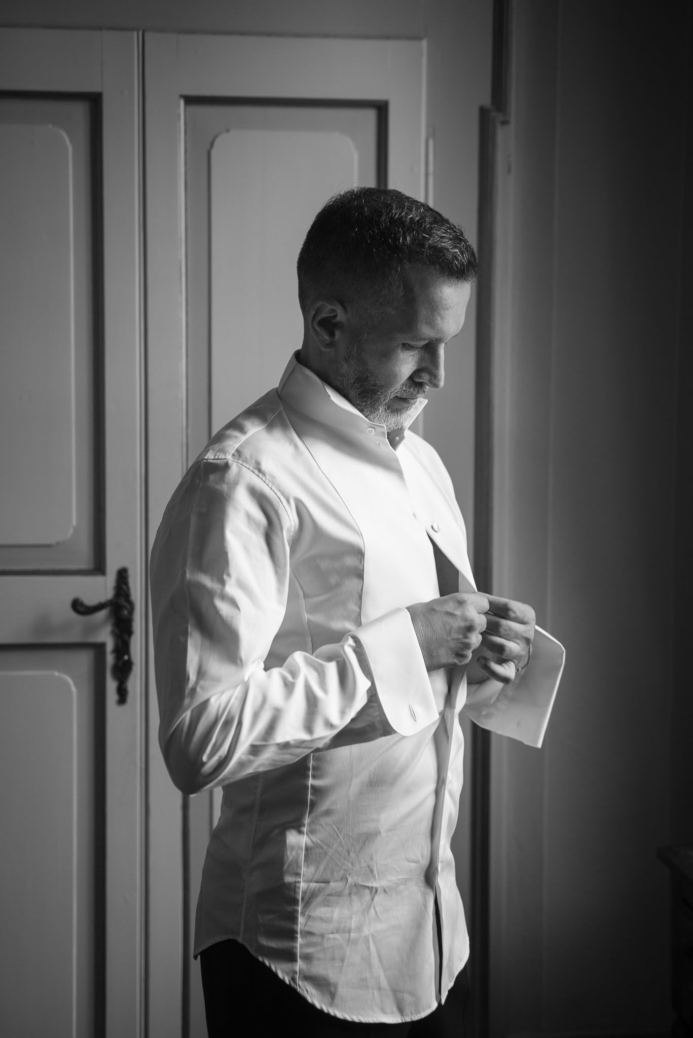 Side profile of someone buttoning up a crisp white dress shirt in moody lighting.