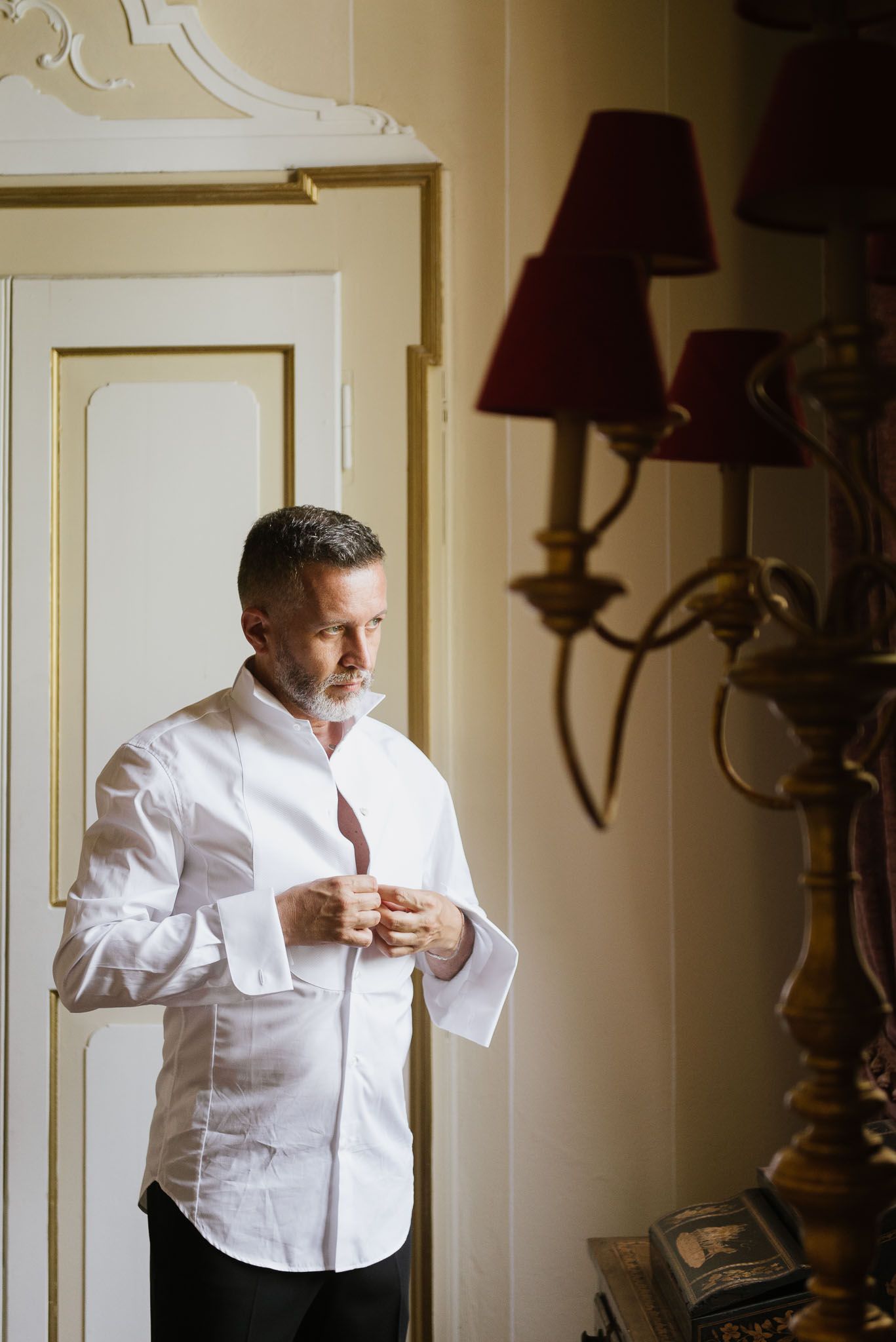 Natural light portrait of someone buttoning a white shirt near an antique lamp.