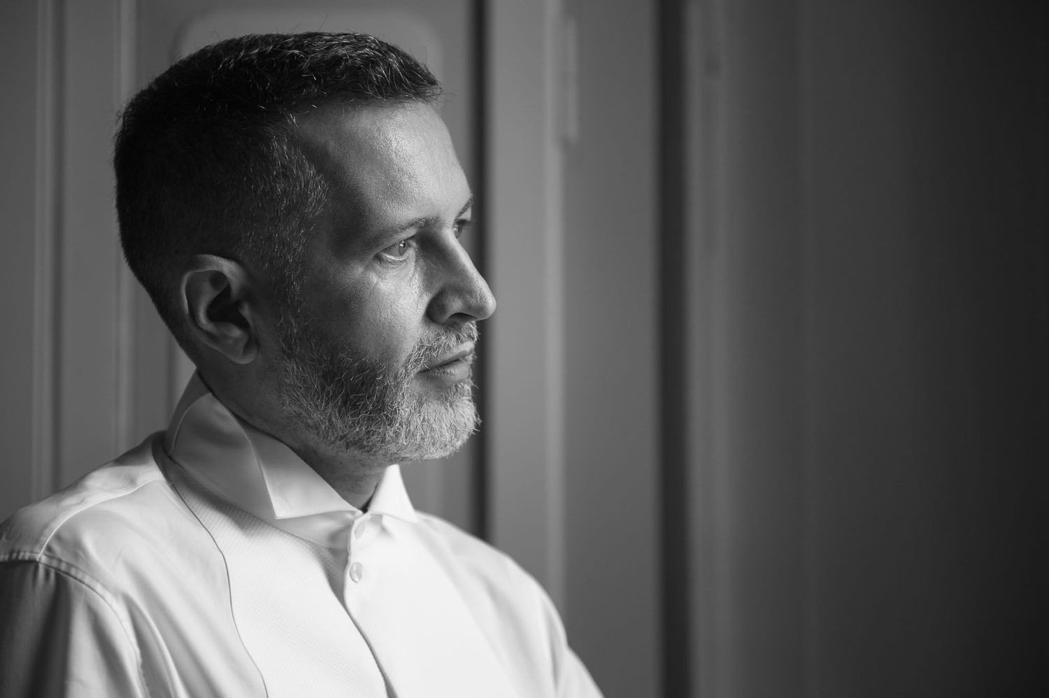 Black and white profile portrait of a well-groomed man in a white shirt gazing thoughtfully by a window.
