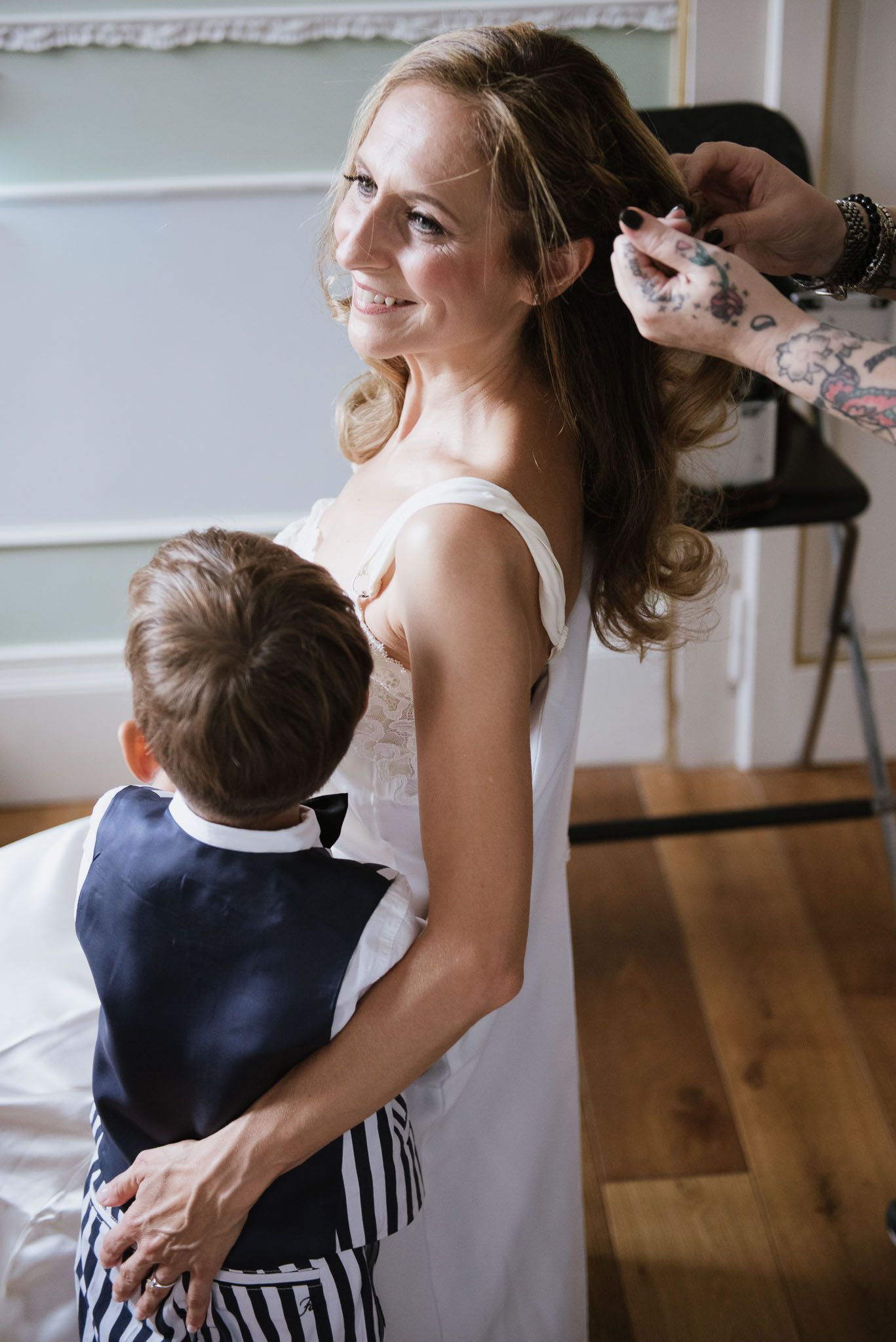Woman in white dress getting hair styled while holding young child in formal wear.