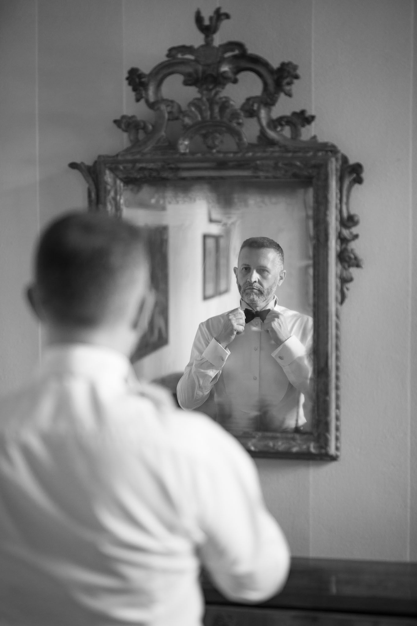 Man in white shirt adjusting bow tie while looking in ornate vintage mirror.