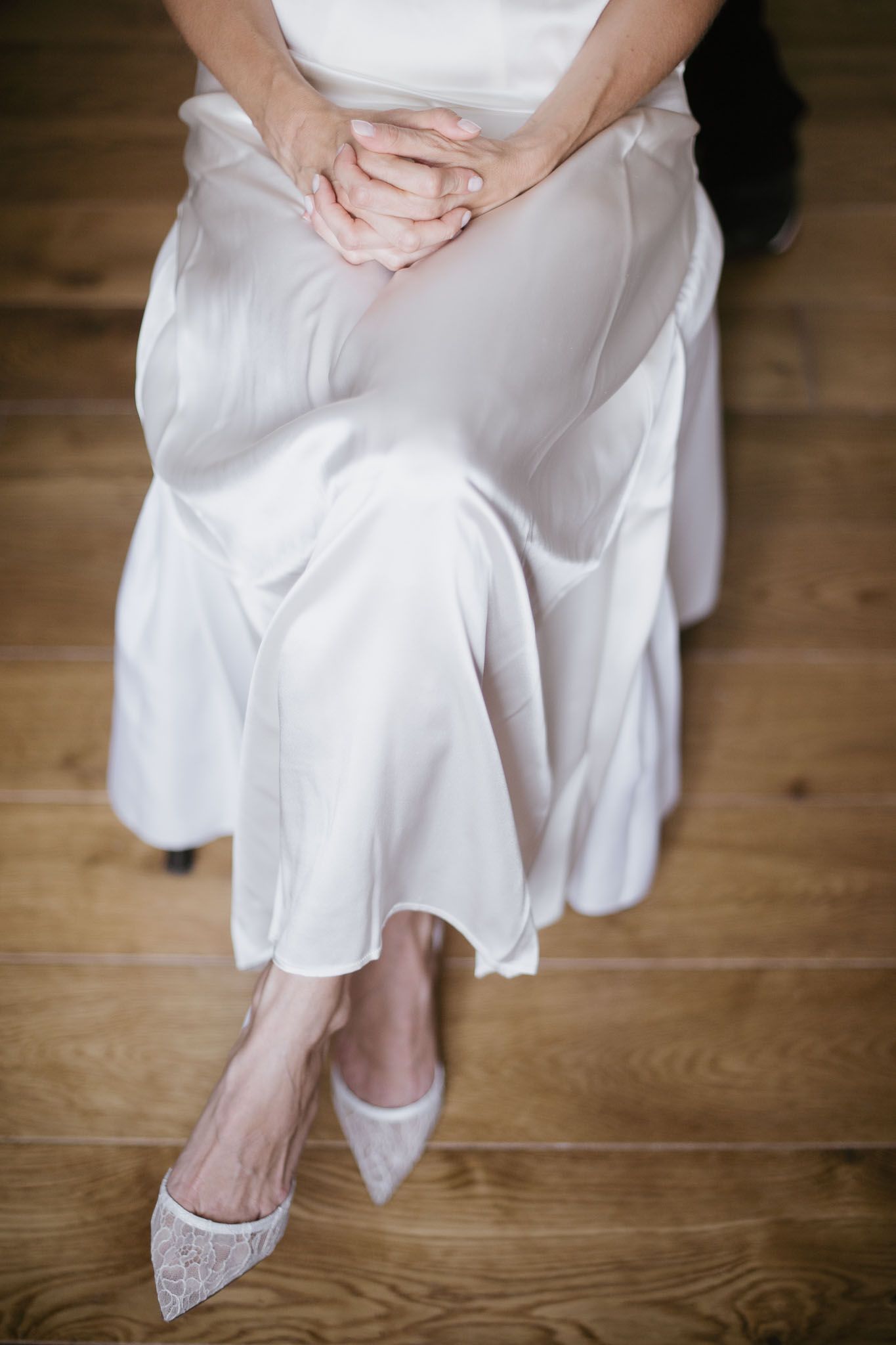 Close-up of white flowing dress and lace shoes on wooden floor with folded hands.