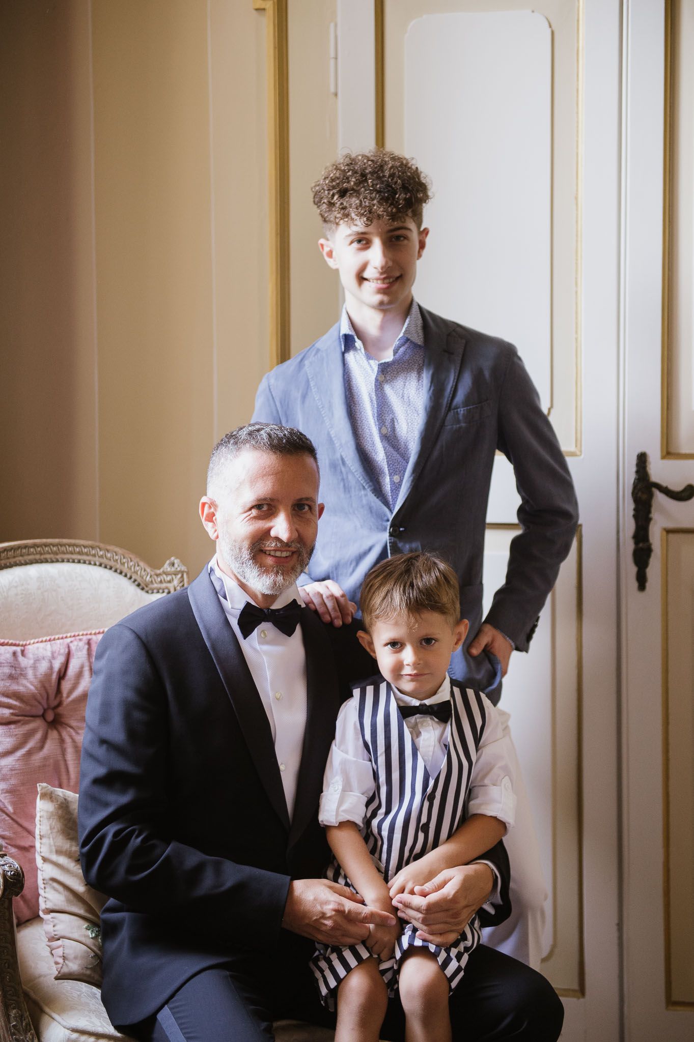 Three people in formal attire pose together near an elegant chair in an indoor setting.