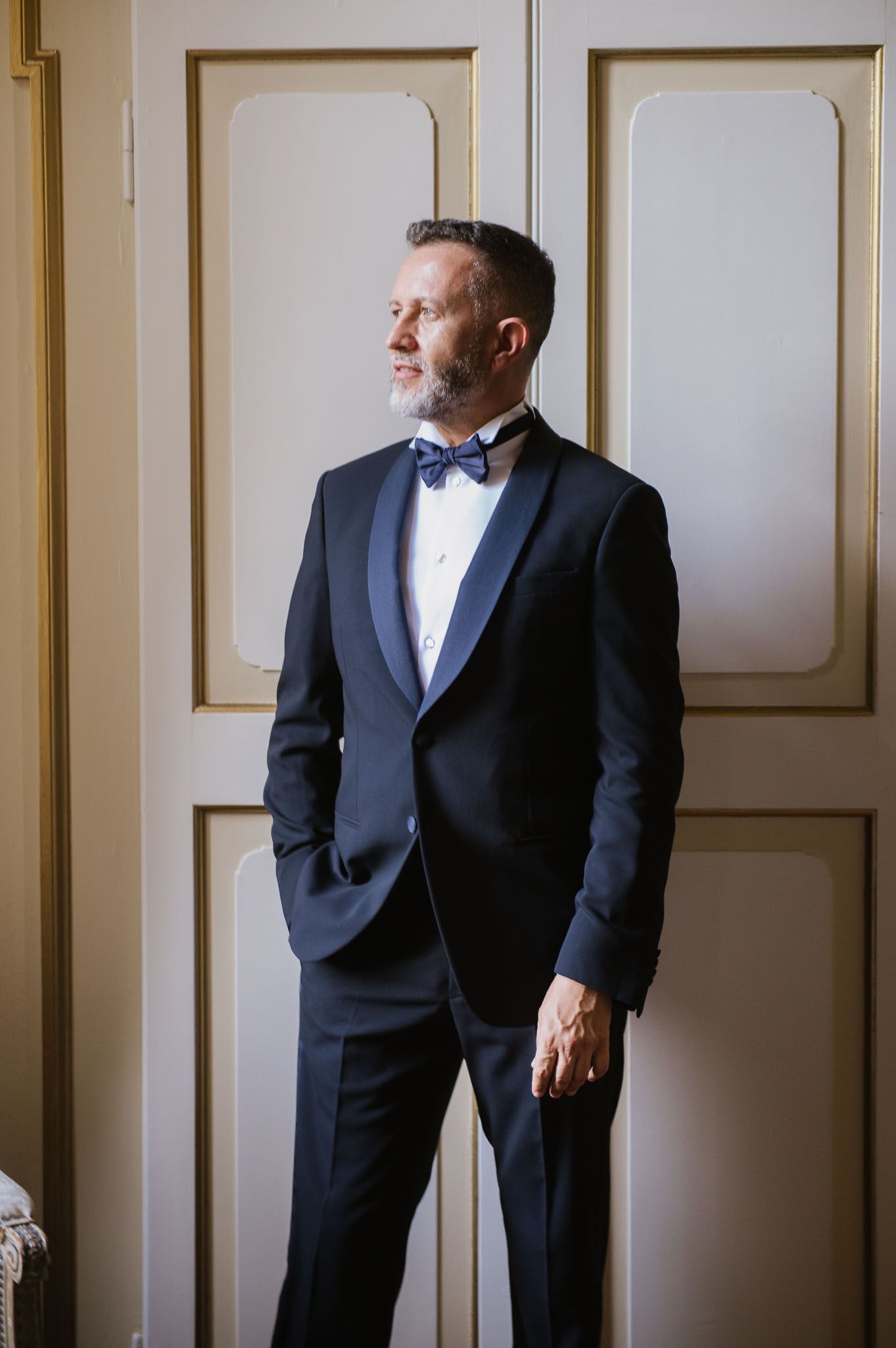 Man in classic tuxedo and bow tie poses confidently against paneled door.