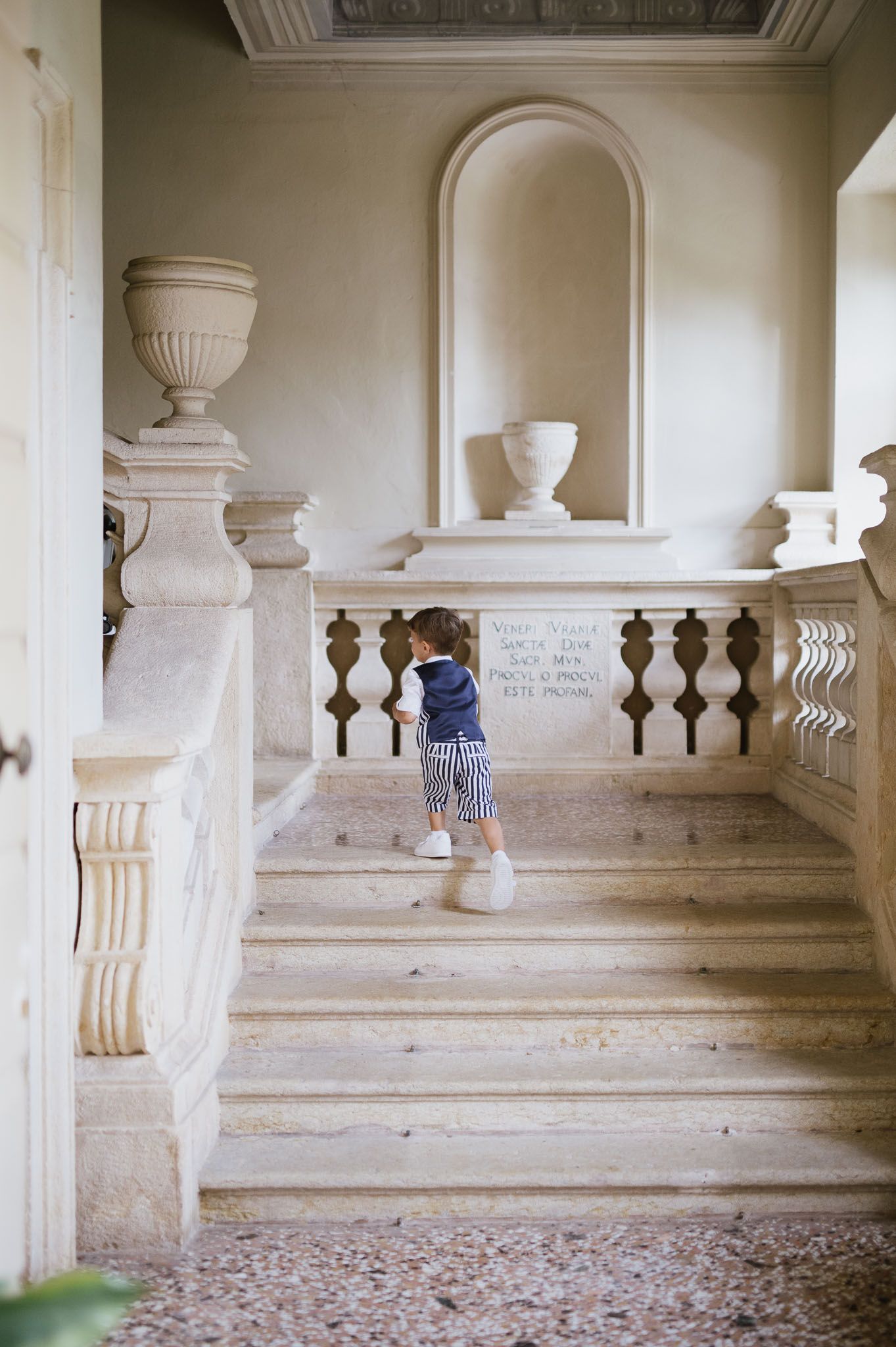 A small child in striped overalls explores ornate marble stairs in a grand building.
