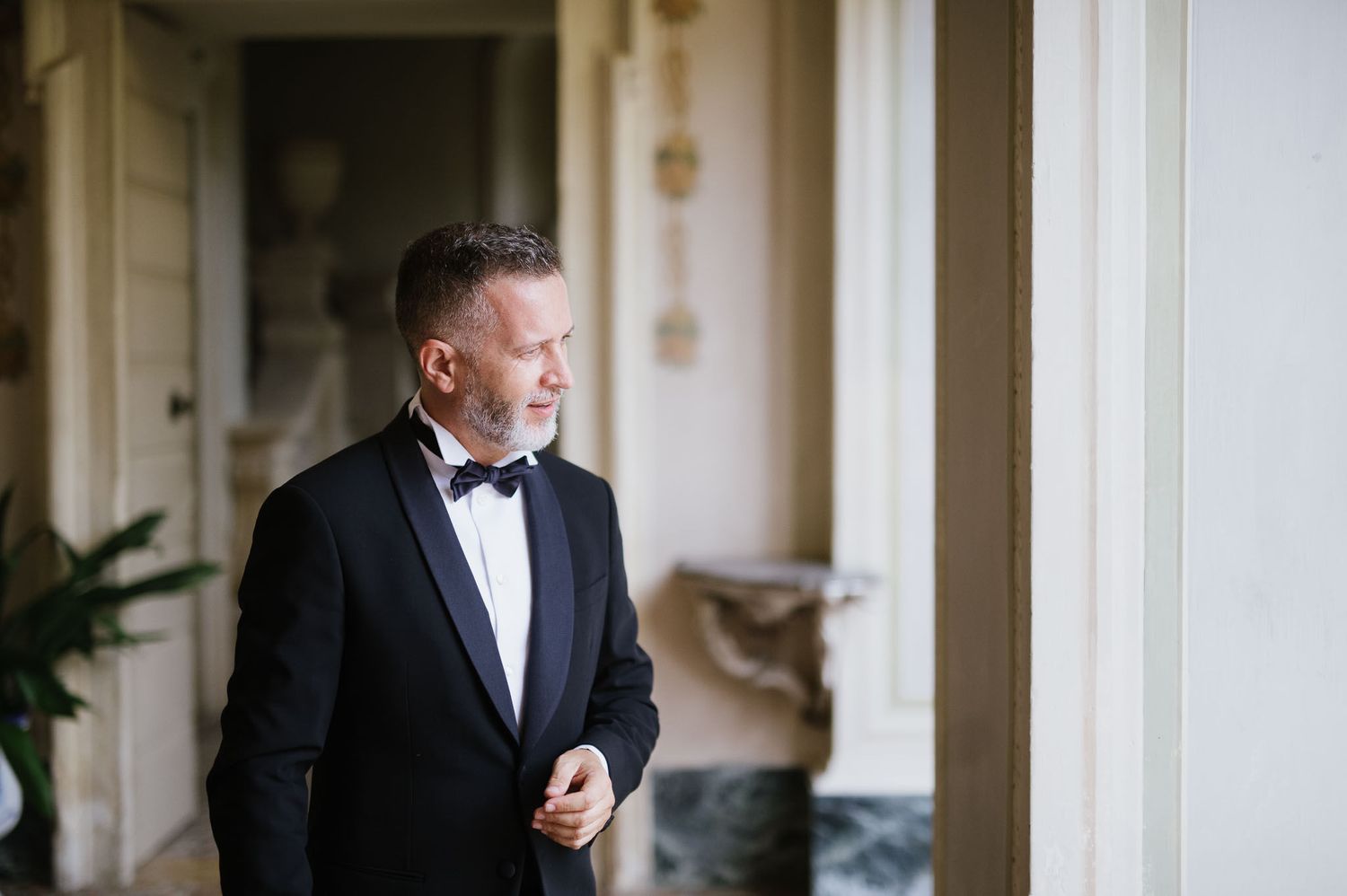 A person in a black tuxedo and bow tie stands by a window in an elegant room.