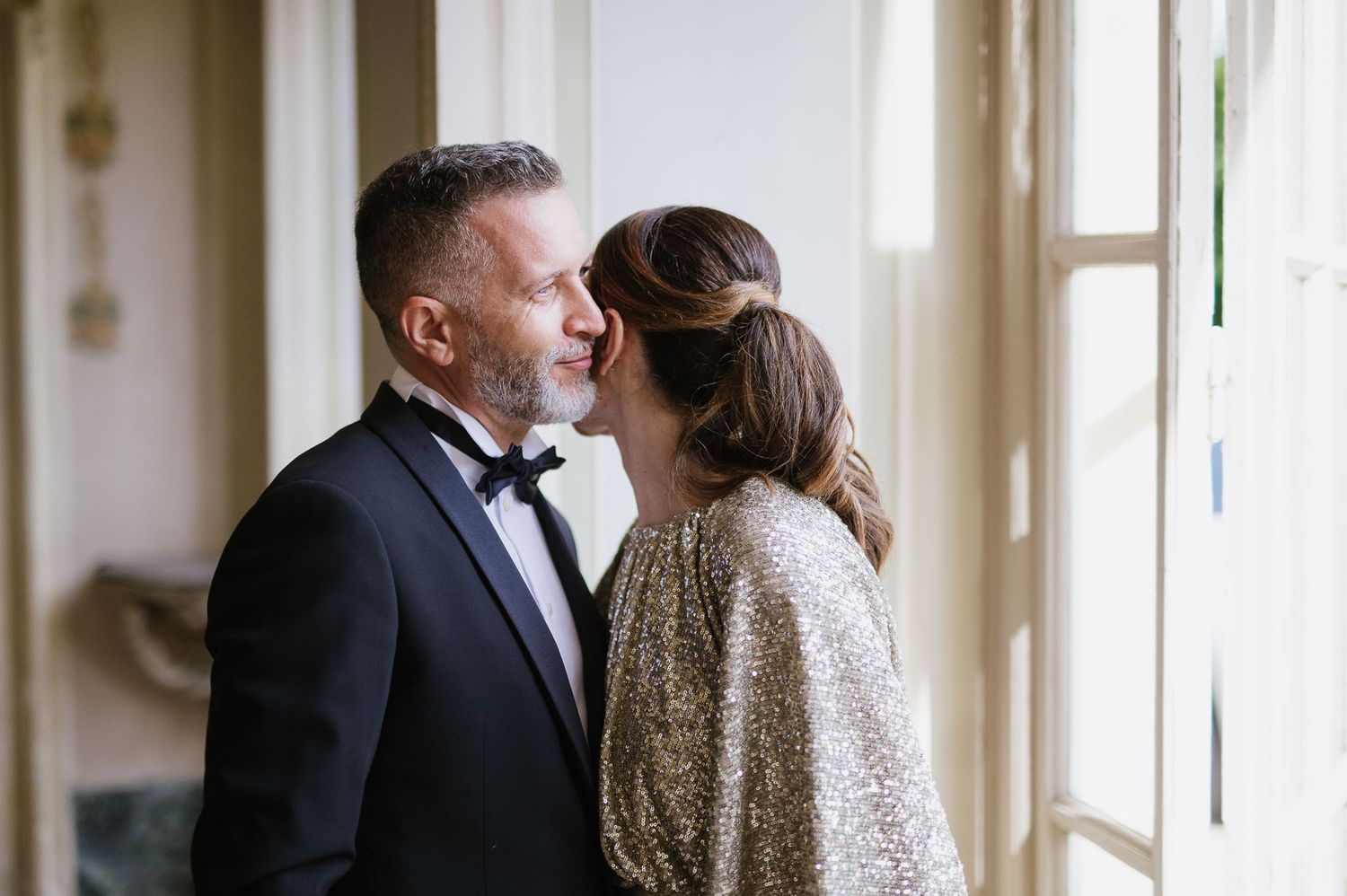 A couple in formal evening wear share an intimate moment by a window.
