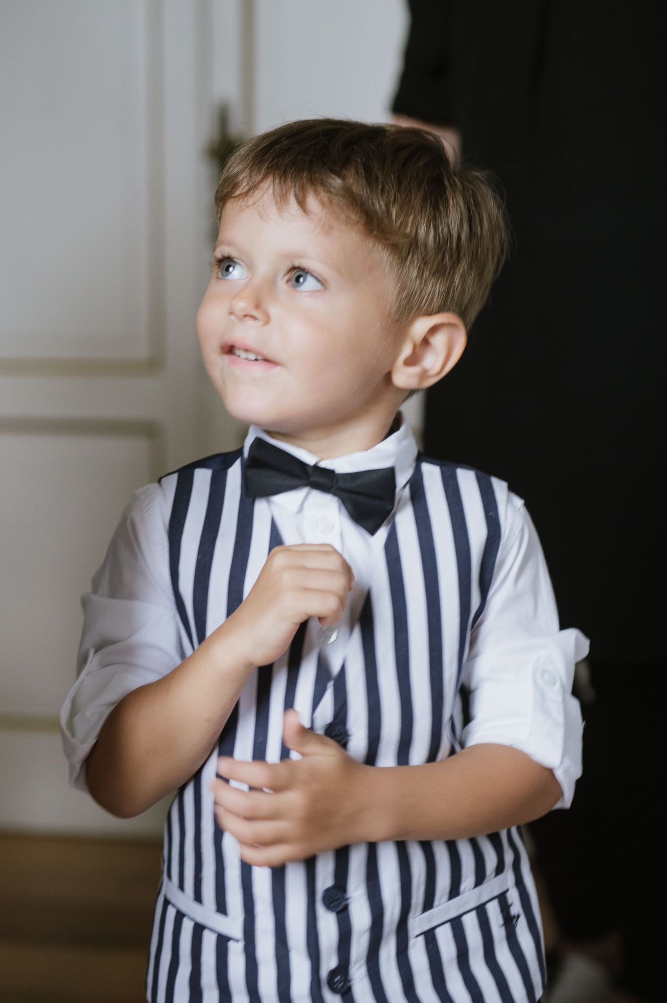 A young child in a striped vest and bow tie looks up while adjusting formal wear.