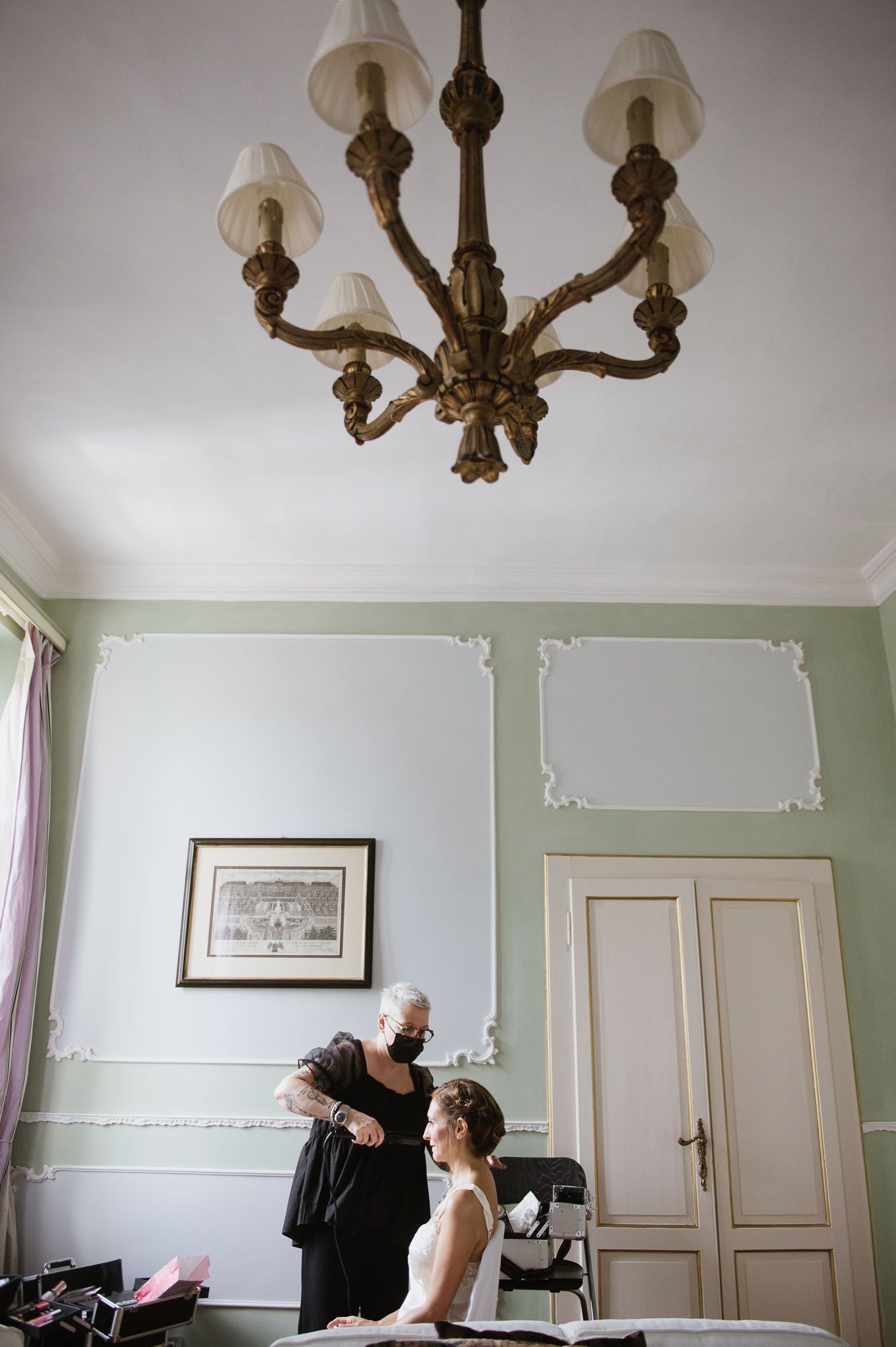 An ornate vintage chandelier hangs above as preparations take place in an elegant room.