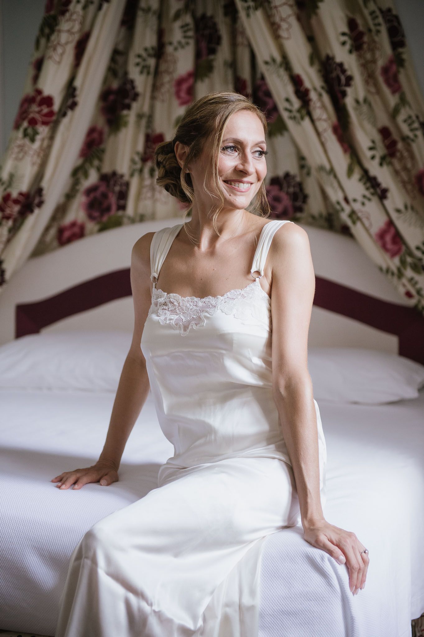 A woman in a white dress sits on a bed with floral curtains in the background.
