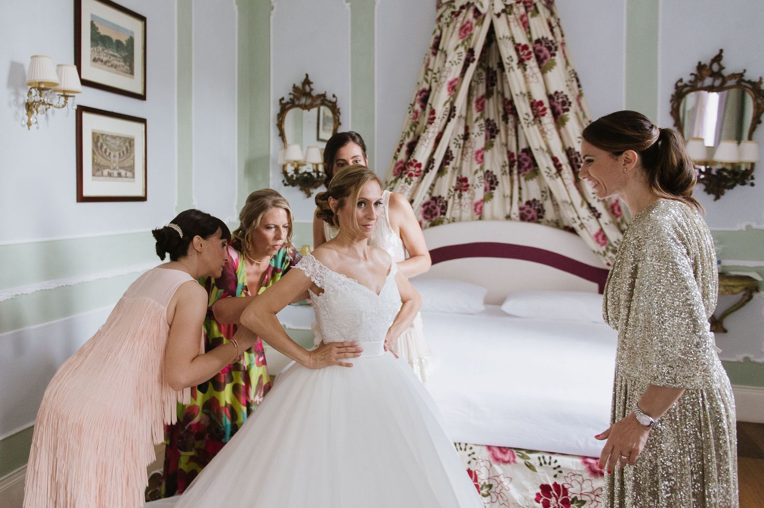 A bridal party helps with wedding dress preparations in an elegant bedroom.