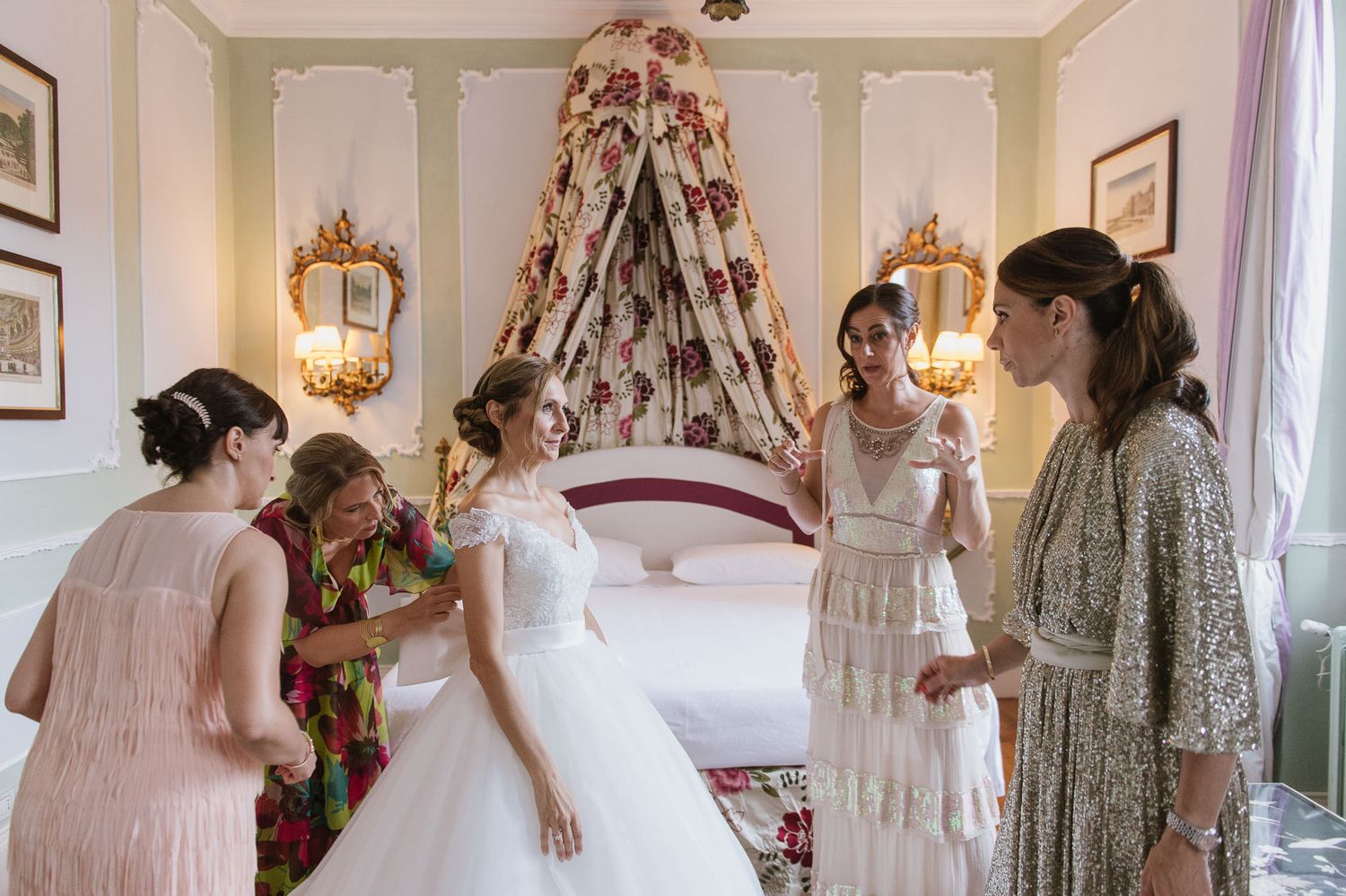 Wedding party helps bride with final preparations in an elegant bedroom with floral canopy.