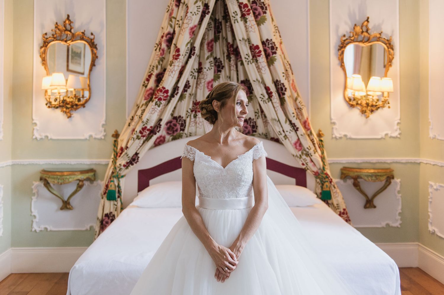 A bride in a classic white gown sits elegantly on a bed beneath ornate wall sconces.