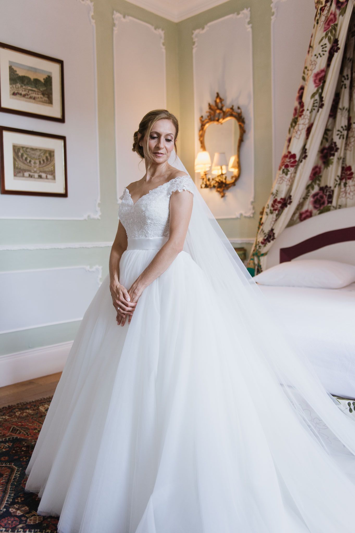 Elegant bride in white off-shoulder ballgown poses in vintage-styled room with ornate mirror.