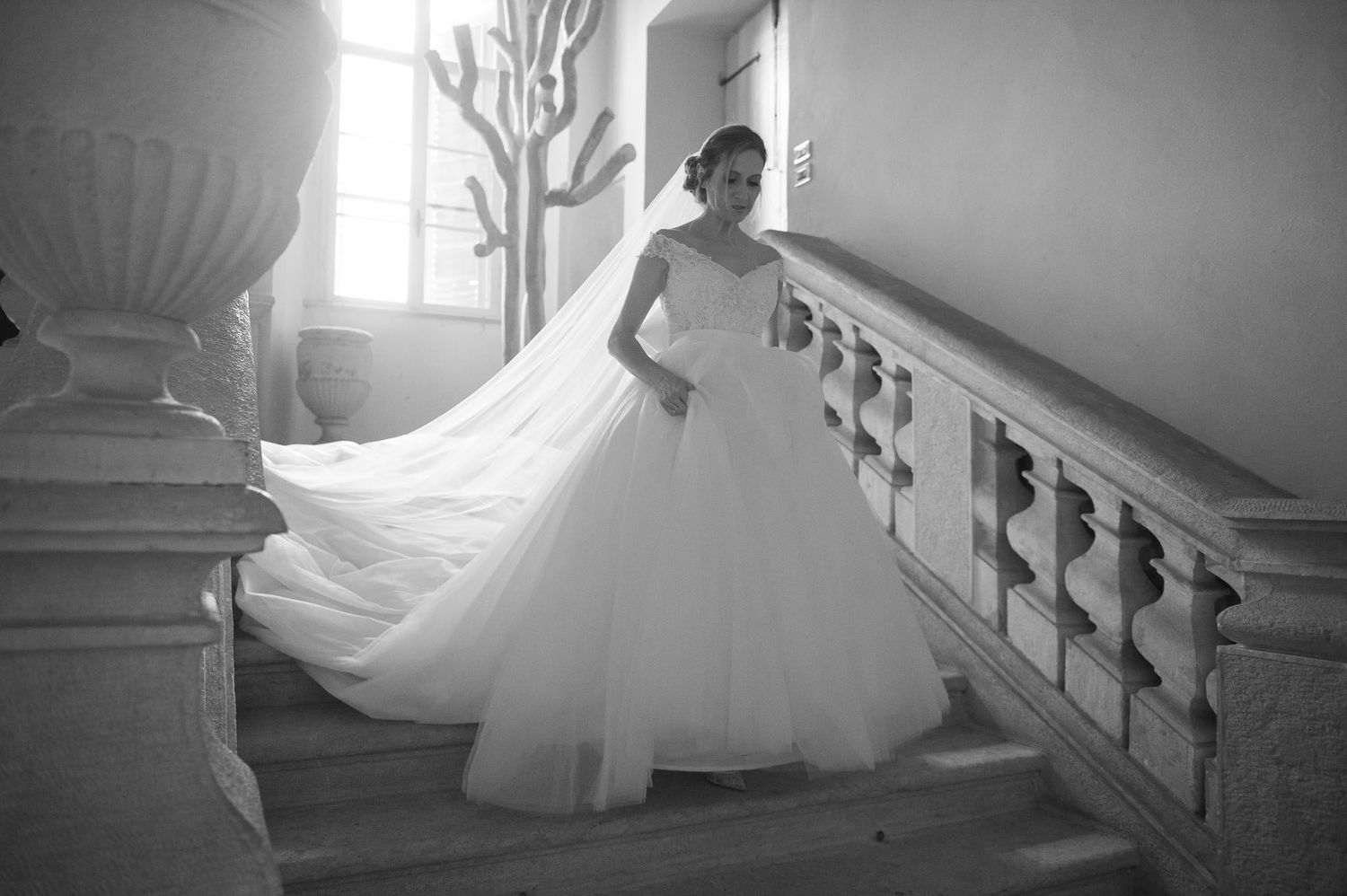 Bride in flowing white gown poses dramatically on grand staircase in black and white.