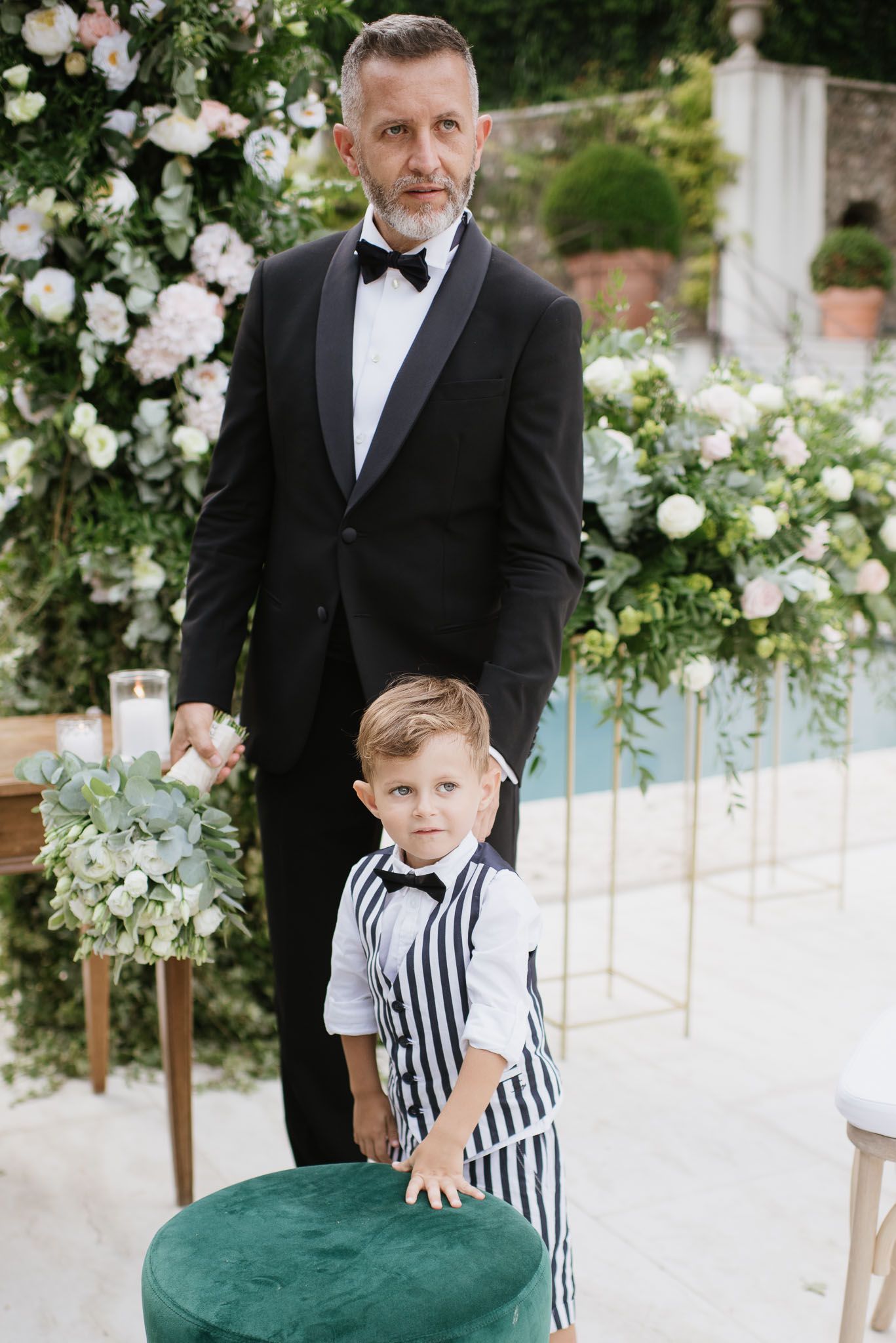Groom in black tuxedo stands with young boy in striped vest near floral arch.