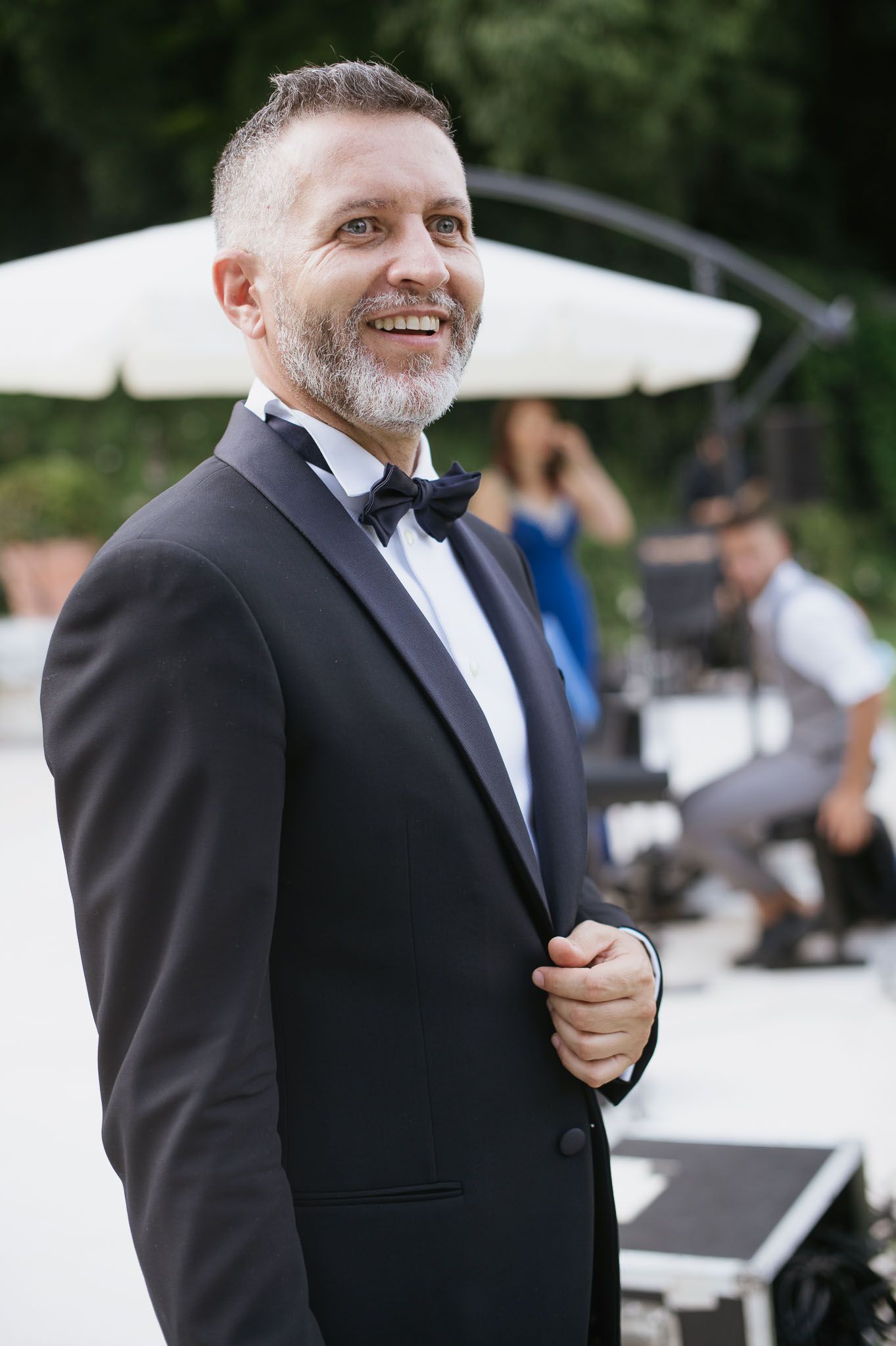Groom smiling in classic black tuxedo and bowtie at outdoor wedding venue.