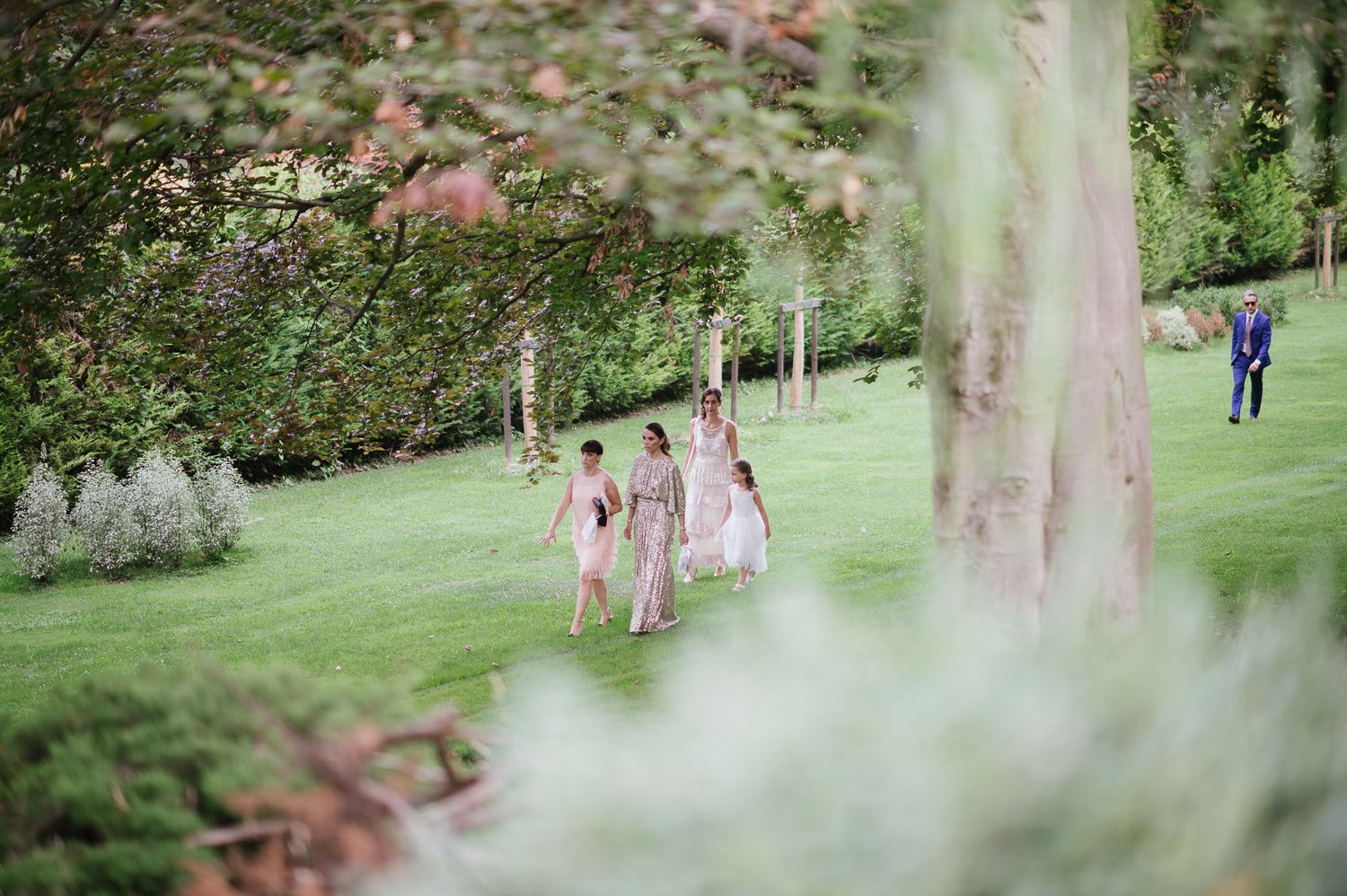 Wedding party members in pastel dresses walk across a lush green lawn at an outdoor celebration.