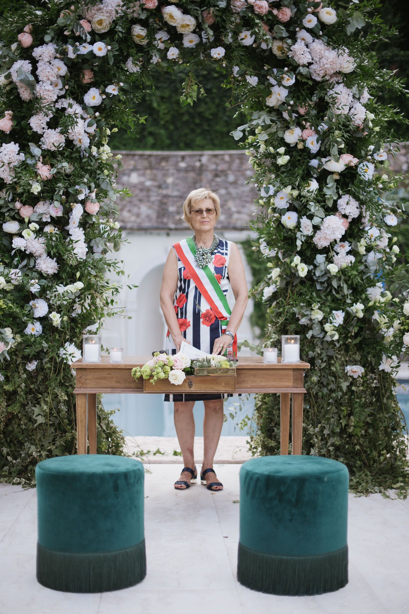 Elegant floral arch with pink and white roses frames an outdoor ceremony setup with emerald velvet stools.