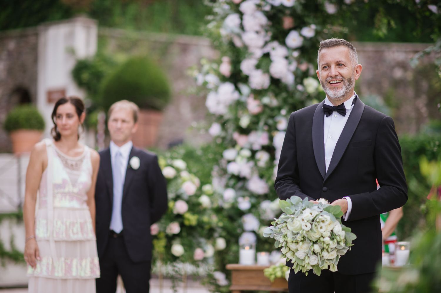 Classic wedding ceremony scene with floral arch and formal attire against stone wall backdrop.