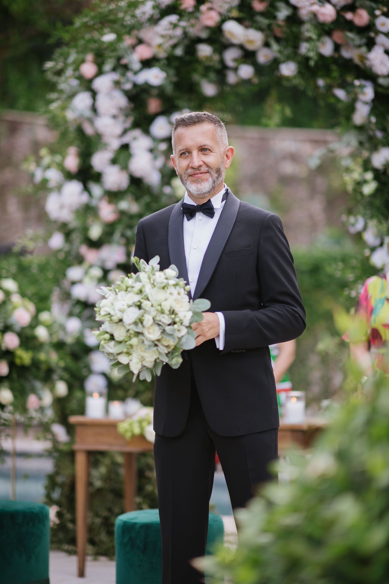 Formal wedding attire against romantic floral arch backdrop with white bouquet.