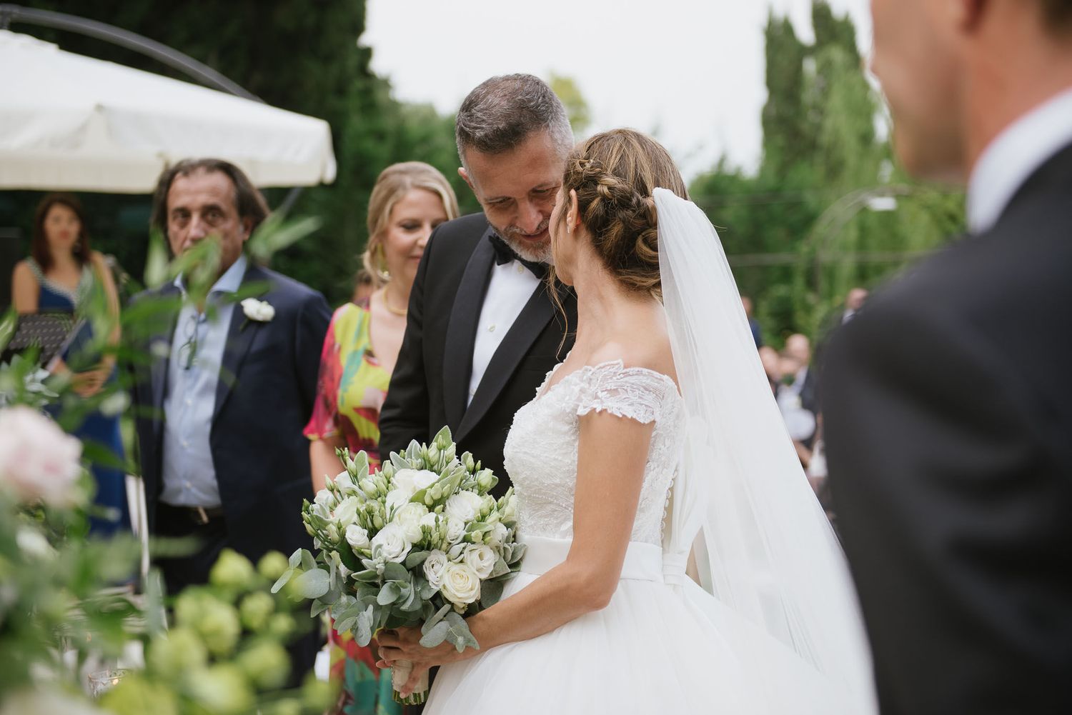 A romantic outdoor wedding ceremony moment with white floral bouquets and elegant attire against a lush garden backdrop.