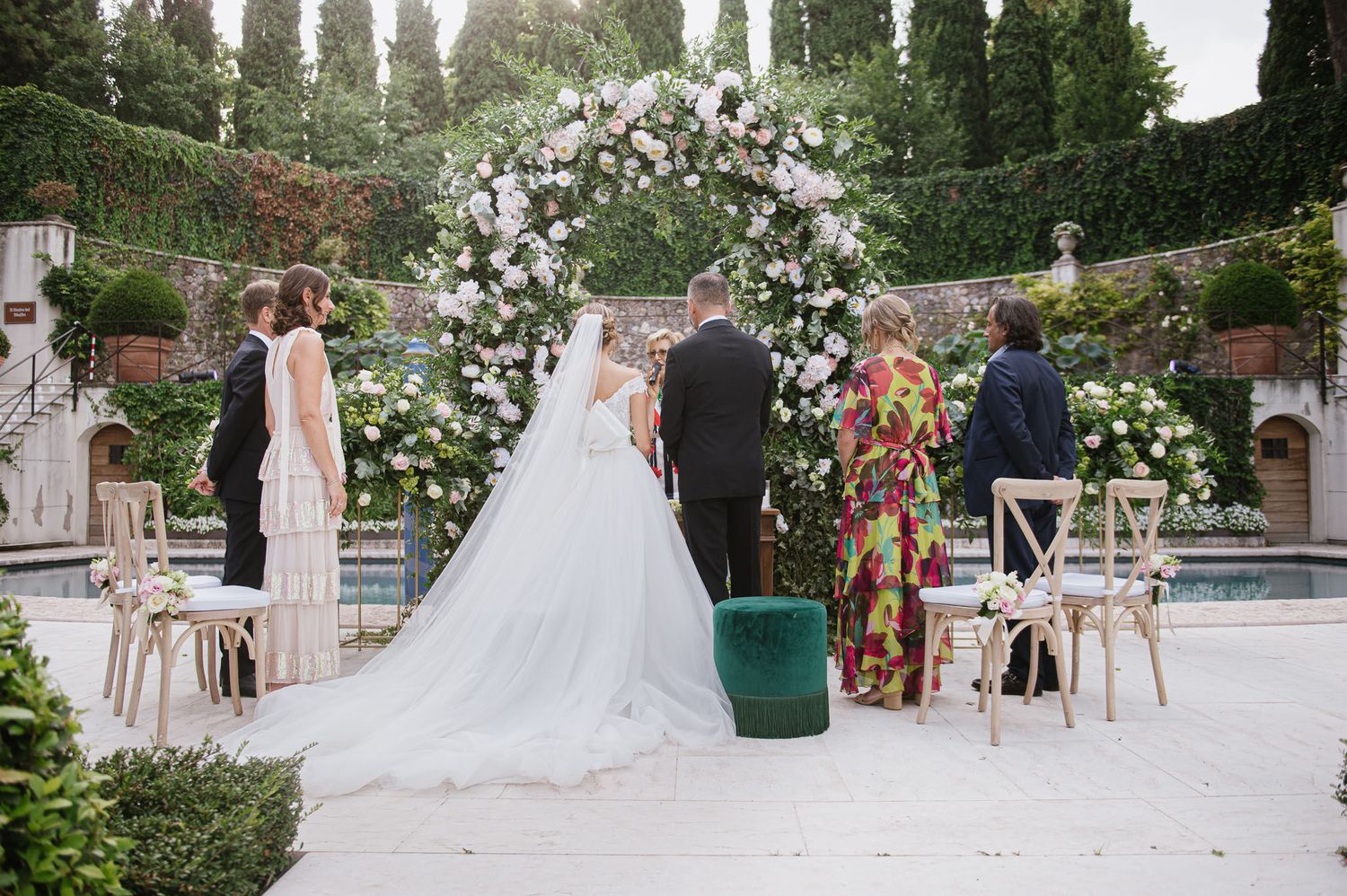An elegant wedding ceremony under a floral arch with white chairs and cypress trees in the background.
