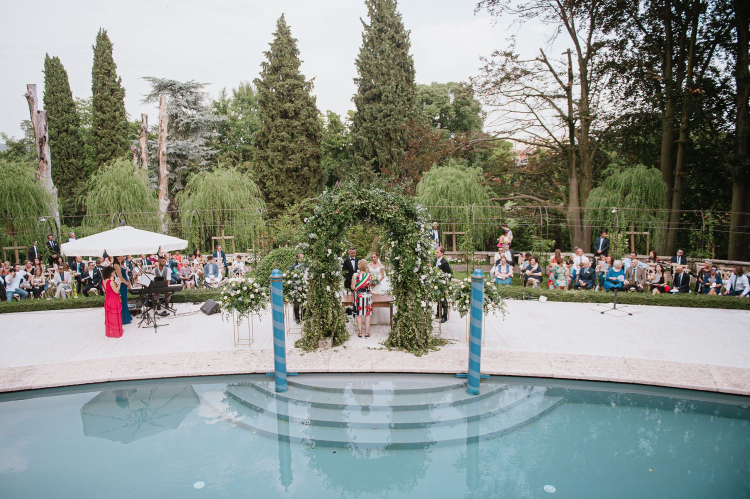 A wedding ceremony setup near a pool with a floral arch and cypress trees creating a stunning backdrop.