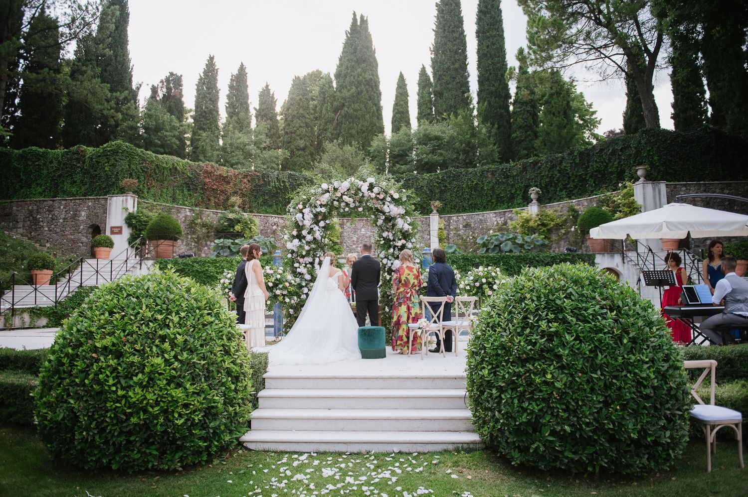A scenic garden wedding setup with a floral arch, manicured bushes, and cypress trees creating a romantic atmosphere.