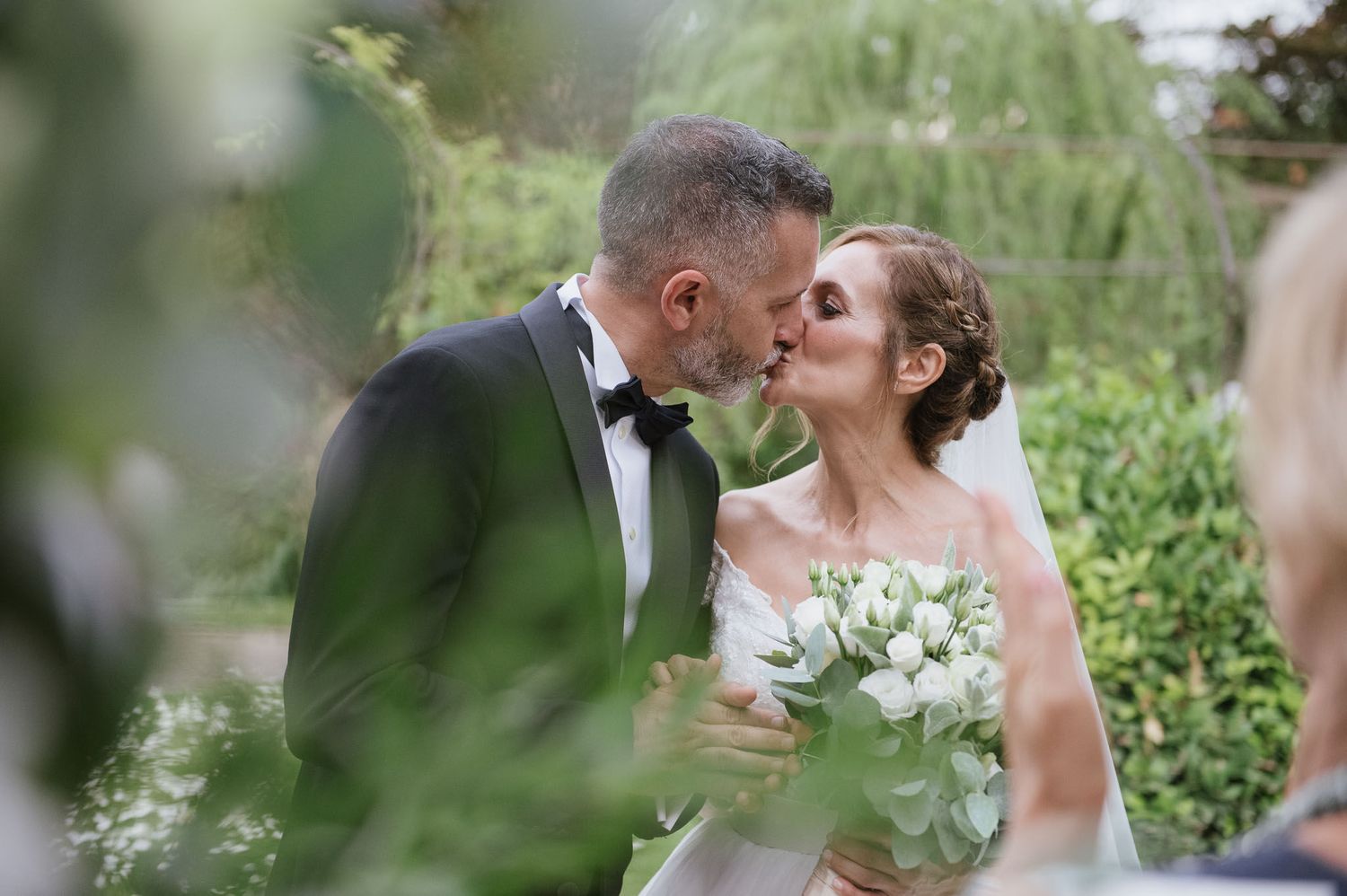 A tender wedding moment captured through green foliage in an outdoor garden setting.