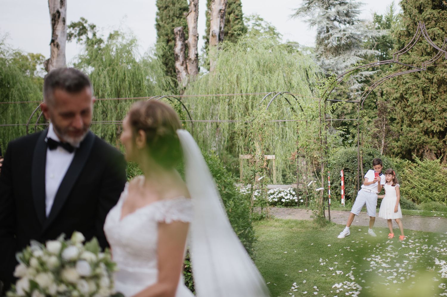 Bride and groom walking under floral arch in elegant garden setting with cypress trees.