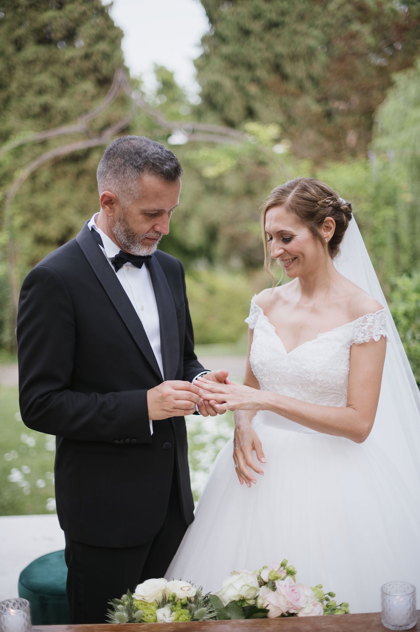 A touching ring exchange moment during an outdoor wedding ceremony with white floral decorations.