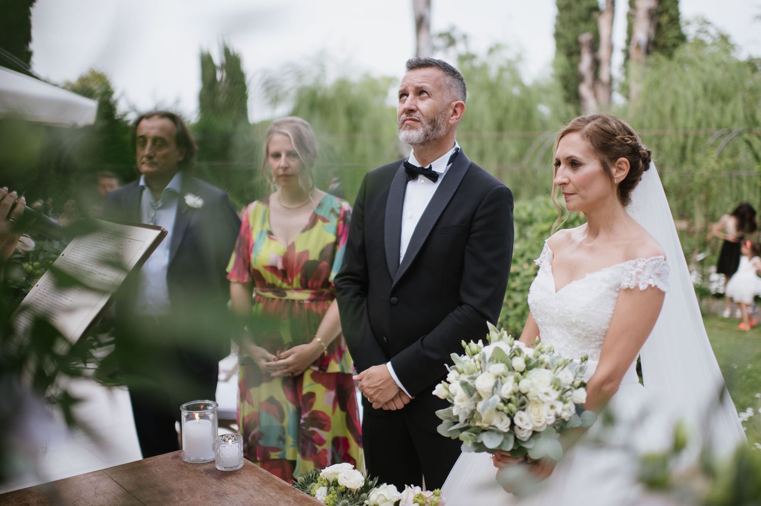 A wedding ceremony takes place outdoors with guests standing amid greenery and formal attire.