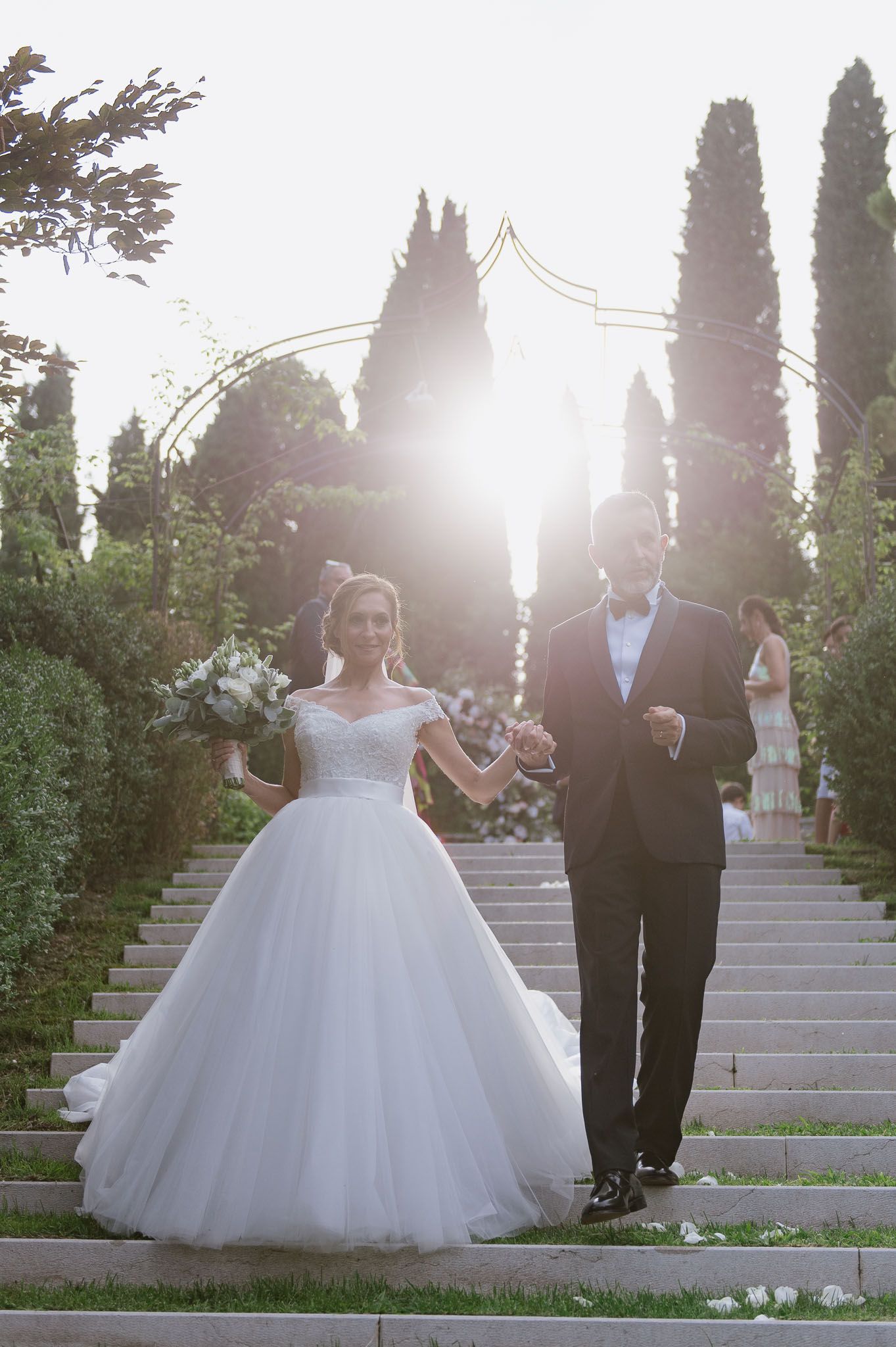 A dreamy backlit wedding portrait on stone steps with cypress trees creating a magical atmosphere.