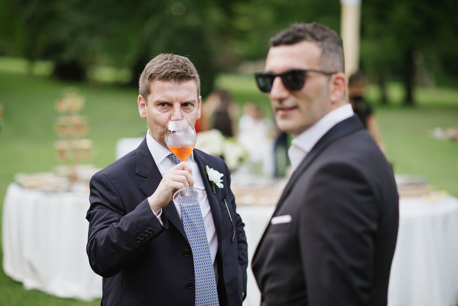 Two groomsmen in suits enjoying cocktails during an outdoor garden reception.