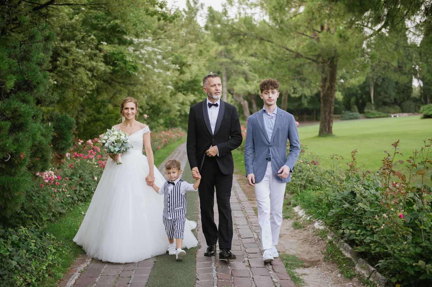 Wedding party walking along a garden path lined with roses and greenery on a sunny day.
