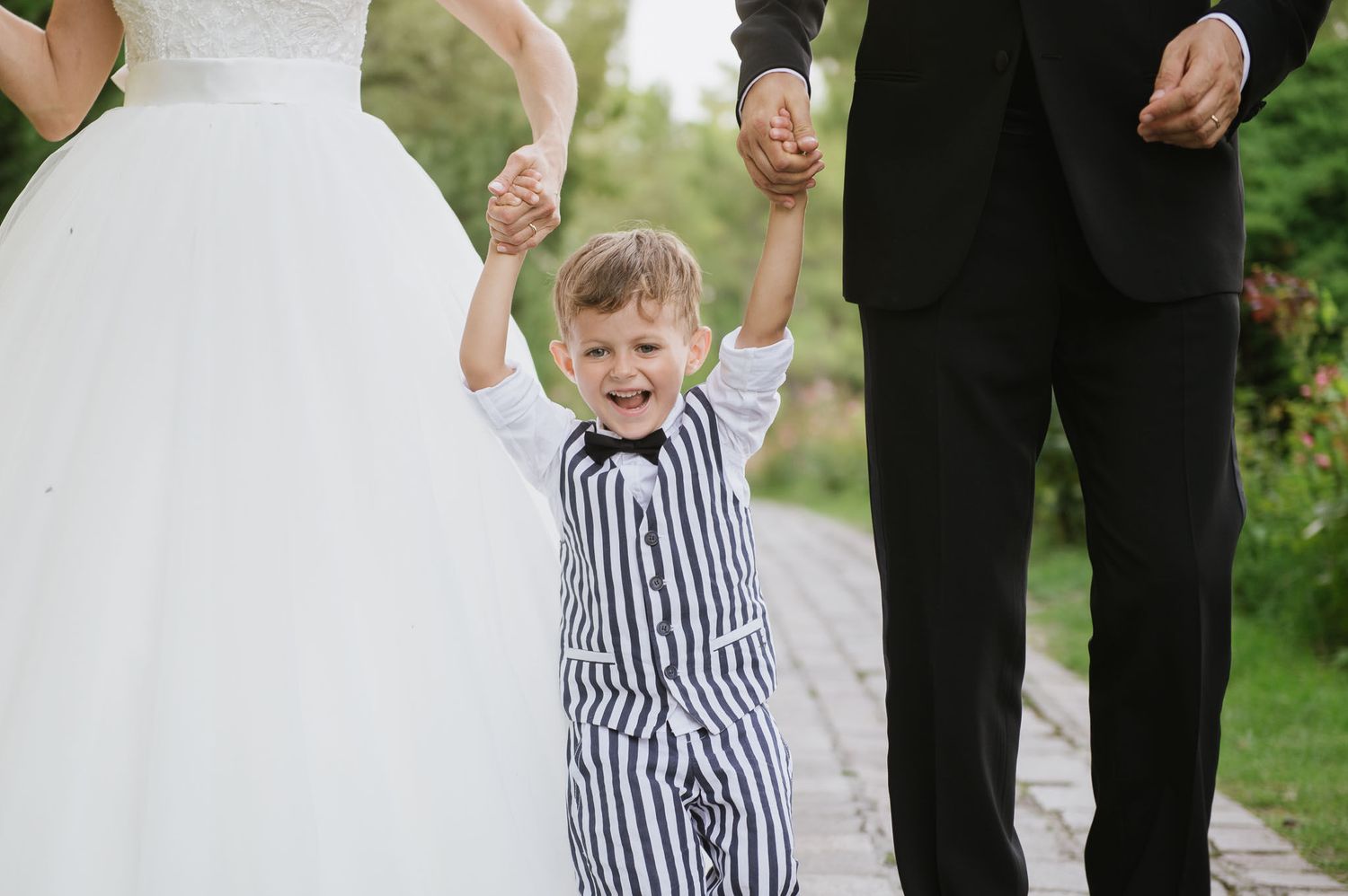 Young boy in striped outfit joyfully holding hands with adults in wedding attire.