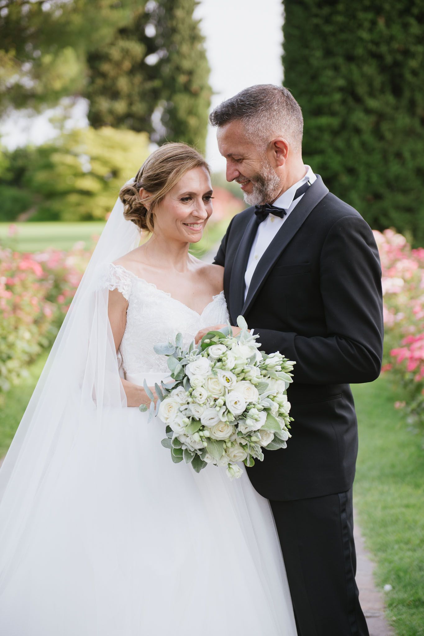 Elegant couple sharing tender moment in garden with white bouquet and classic attire.